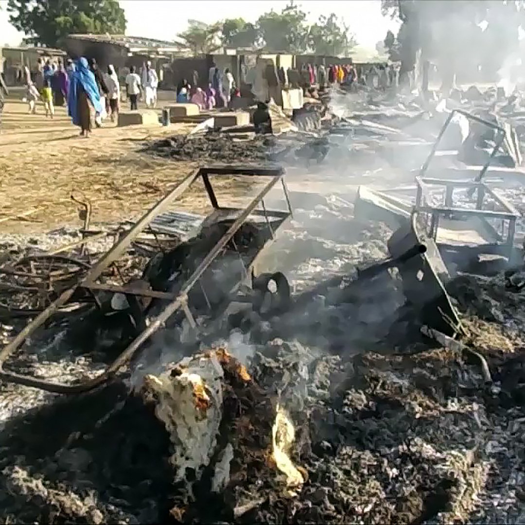 L'image montre une scène de destruction, où des débris brûlés et des objets en métal tordu sont dispersés sur le sol. De la fumée s'élève de restes calcinés, suggérant qu'un incendie a eu lieu récemment. À l'arrière-plan, on aperçoit des personnes en train de se déplacer, sur un terrain qui semble être une communauté ou un marché. L'atmosphère est sombre et le paysage est marqué par les dégâts causés par le feu.