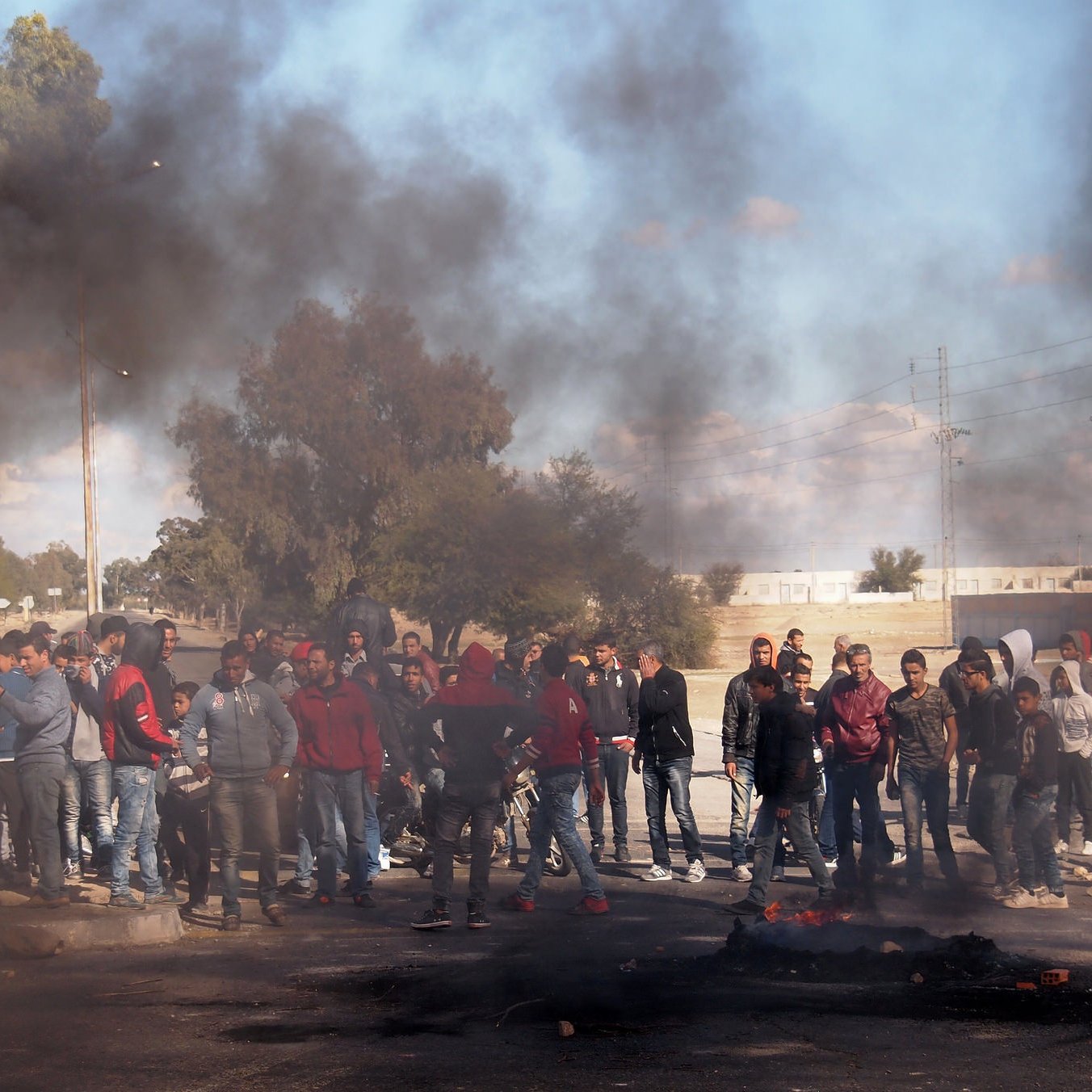 L'image montre un groupe de personnes rassemblées sur une route où de la fumée noire s'élève, probablement à cause de pneus ou d'autres matériaux en feu. Les manifestants semblent préoccupés et engagés dans un rassemblement, tandis que l'arrière-plan indique un paysage urbain avec des arbres et des infrastructures. La scène évoque une atmosphère de tension ou de protestation.