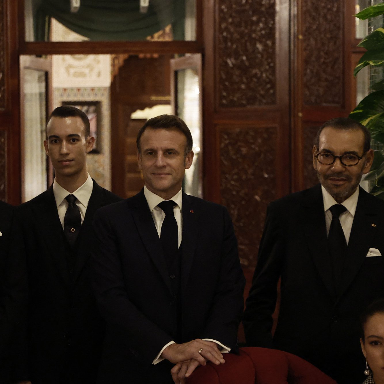 يظهر في الصورة مجموعة من الرجال يرتدون بدلات رسمية سوداء. يقع شخص في الوسط يبدو أنه في موقع قيادي أو مهم، حيث يرتدي بدلة داكنة وربطة عنق. إلى جانبه، هناك شخص آخر يرتدي أيضًا بدلة، بالإضافة إلى رجلين آخرين على الجانبين. الخلفية تتضمن تفاصيل معمارية جميلة، مع نقوش وزخارف تقليدية. هناك نباتات خضراء تضيف لمسة طبيعية إلى المشهد. 분위기 يظهر احتفاليًا ورسميًا.