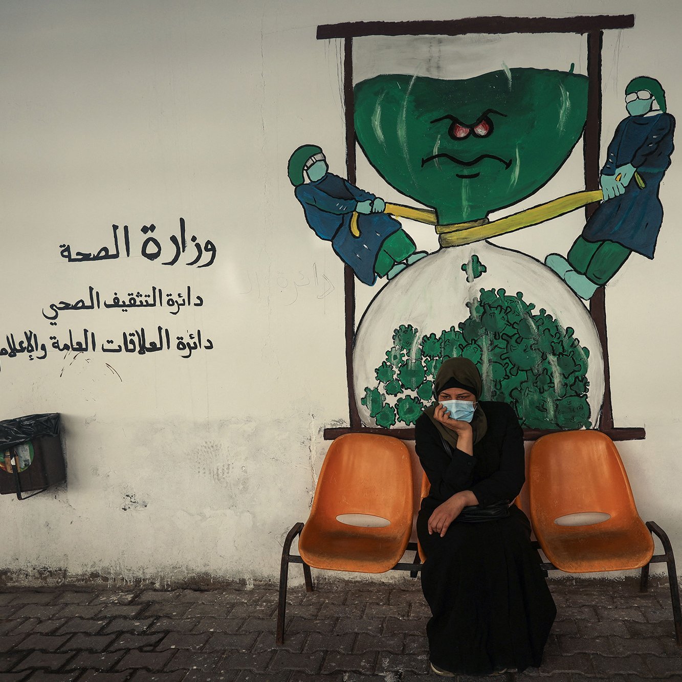 الصورة تظهر امرأة جالسة على كرسيين في منطقة تبدو وكأنها مركز صحي أو مستشفى. تحمل المرأة ملامح الجدية، وتضع كمامة على وجهها. خلفها يوجد جدار م painted يوضح رسماً يجسد فكرة وزارة الصحة، حيث يظهر شخصيتان ترتديان بزة وقاية، تسعيان لشد أو دفع شيء كبير يشبه الخضار أو الملفوف. الألوان المستخدمة في الجدار تعكس موضوع الصحة، مع تركيبات واضحة ورموز مثل القفازات والكمامات.