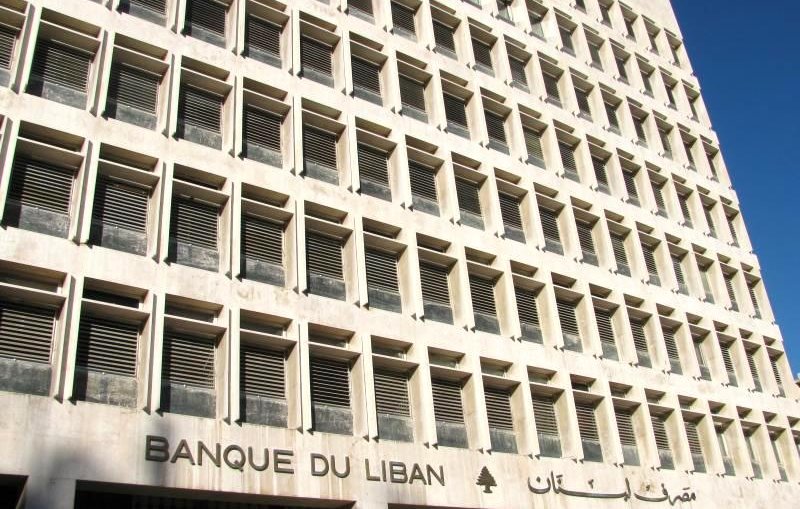 L'image montre un bâtiment moderne qui abrite la Banque du Liban. La façade est caractérisée par un design géométrique, avec de nombreuses fenêtres disposées en grille, donnant une impression de symétrie. On peut également voir le nom de la banque inscrit en arabe et en français, ainsi qu'un symbole représentant un cèdre, un arbre emblématique du Liban. L'architecture est sobre et imposante, typique des institutions financières. Le ciel est clair, suggérant une journée ensoleillée.