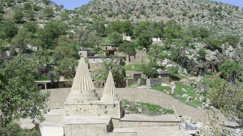 L'image montre un paysage montagneux avec une structure architecturale distinctive au centre. Cette structure présente deux tours ou pinacles coniques, typiques d'un style architectural particulier. Autour, on aperçoit des bâtiments en pierre et en béton, ainsi que des arbres verdoyants qui ajoutent une touche de nature. Le terrain est légèrement accidenté, avec des collines à l'arrière-plan, créant un environnement pittoresque et tranquille.