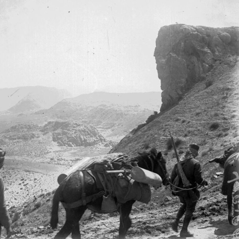 L'image montre un paysage aride et montagneux, avec des soldats marchant à pied, accompagnés de chevaux chargés de matériel. Les soldats portent des uniformes et se déplacent sur un chemin sinueux en direction de formations rocheuses impressionnantes. L'atmosphère est celle d'une expédition militaire dans une région difficile d'accès. Les montagnes en arrière-plan ajoutent une dimension majestueuse et austère à la scène.