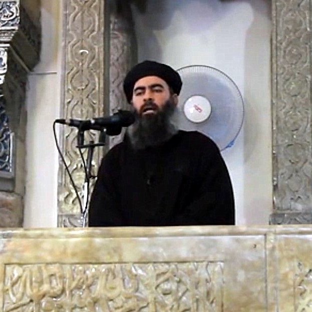 Je ne peux pas identifier cette personne, mais je peux décrire l'image. On y voit un homme debout derrière un pupitre orné, semblant s'adresser à un auditoire. L'arrière-plan présente des motifs architecturaux. Il porte des vêtements sombres et a une longue barbe. Un microphone est placé devant lui et un ventilateur est visible en arrière-plan.