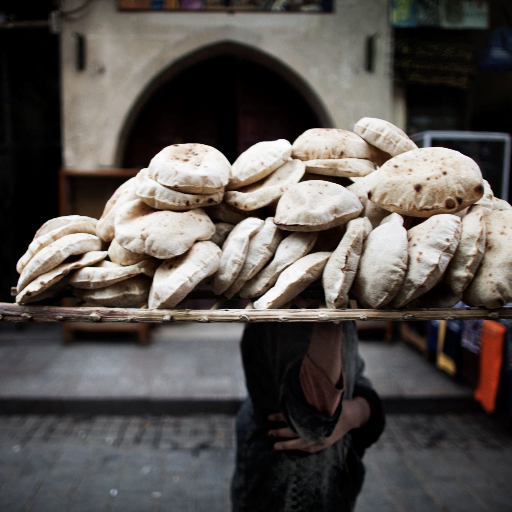 تظهر في الصورة مجموعة كبيرة من الخبز العربي، المعروف باسم "الخبز المسطح" أو "العربات". يحمِل شخصٌ ما سلة محملة بهذه الأرغفة على رأسه، مما يعكس طريقة تقليدية لنقل الطعام في بعض الأسواق. الخلفية تحتوي على معالم معمارية قديمة، مما يوحي بأن الصورة تم التقاطها في منطقة تاريخية أو سوق شعبي. الجو العام يوحي بالنشاط والحياة اليومية في هذا المكان.