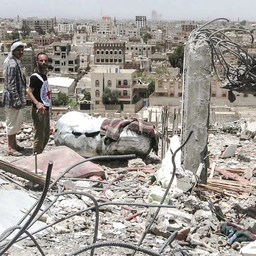 L'image montre deux hommes se tenant au milieu des décombres d'un bâtiment détruit, surplombant une ville en arrière-plan. Les structures environnantes semblent endommagées, avec de nombreux bâtiments partiellement effondrés et une apparence de désolation. Le ciel est clair, mais l'environnement porte des traces évidentes de conflit. Les hommes semblent examine la scène, témoignant d'une situation difficile et dévastatrice.