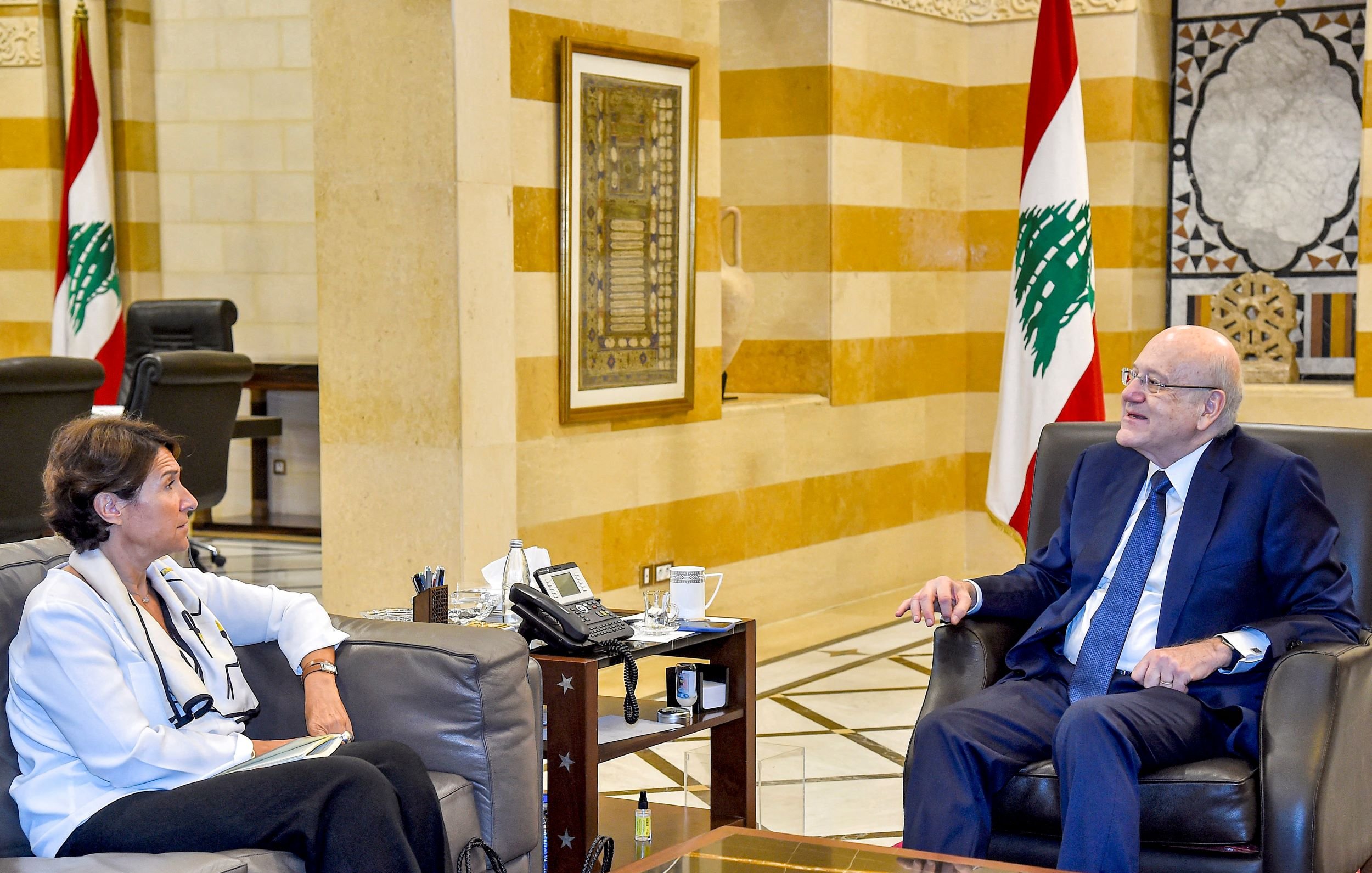 الصورة تظهر اجتماعًا داخل مكتب. يجلس رجل وامرأة على أريكتين. الرجل يبدو في منتصف العمر ويرتدي بدلة رسمية. المرأة ترتدي ملابس عادية، ويبدو أنها تشارك في الحوار. في الخلفية، تُرى ألوان العلم اللبناني، مما يدل على أن الاجتماع قد يكون ضمن إطار حكومي أو رسمي. الأثاث والتصميم الداخلي يعبّران عن الفخامة.