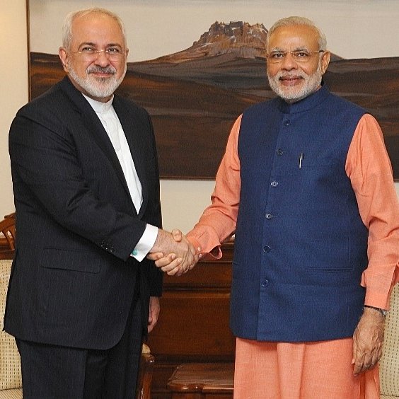 L'image montre deux hommes se serrant la main dans un cadre formel. L'un d'eux porte un costume noir et une chemise blanche, tandis que l'autre est habillé d'une veste bleue et d'une chemise orange. Ils semblent être en train d'échanger une salutation amicale, et l'arrière-plan montre une œuvre d'art représentant un paysage. L'atmosphère semble cordial et professionnelle.