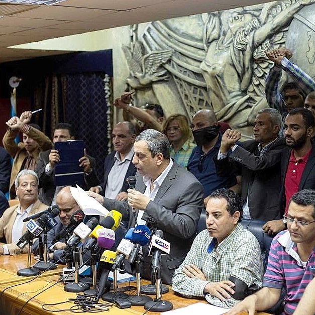 تظهر الصورة اجتماعًا تضامنياً أو مؤتمرًا صحفيًا حيث يجلس عدد من الأشخاص حول طاولة كبيرة، بينما يقف متحدث أمامهم يتحدث إلى الحضور. في الخلفية، يرفع آخرون أيديهم في إشارة إلى الاحتجاج أو التأييد. يظهر البعض في الصورة تفاعلهم مع المتحدث، مما يشير إلى أجواء حماسية. الخلفية تحتوي على منحوتات أو أنماط تزيينية، مما يضيف لمسة رسمية لمكان الاجتماع.