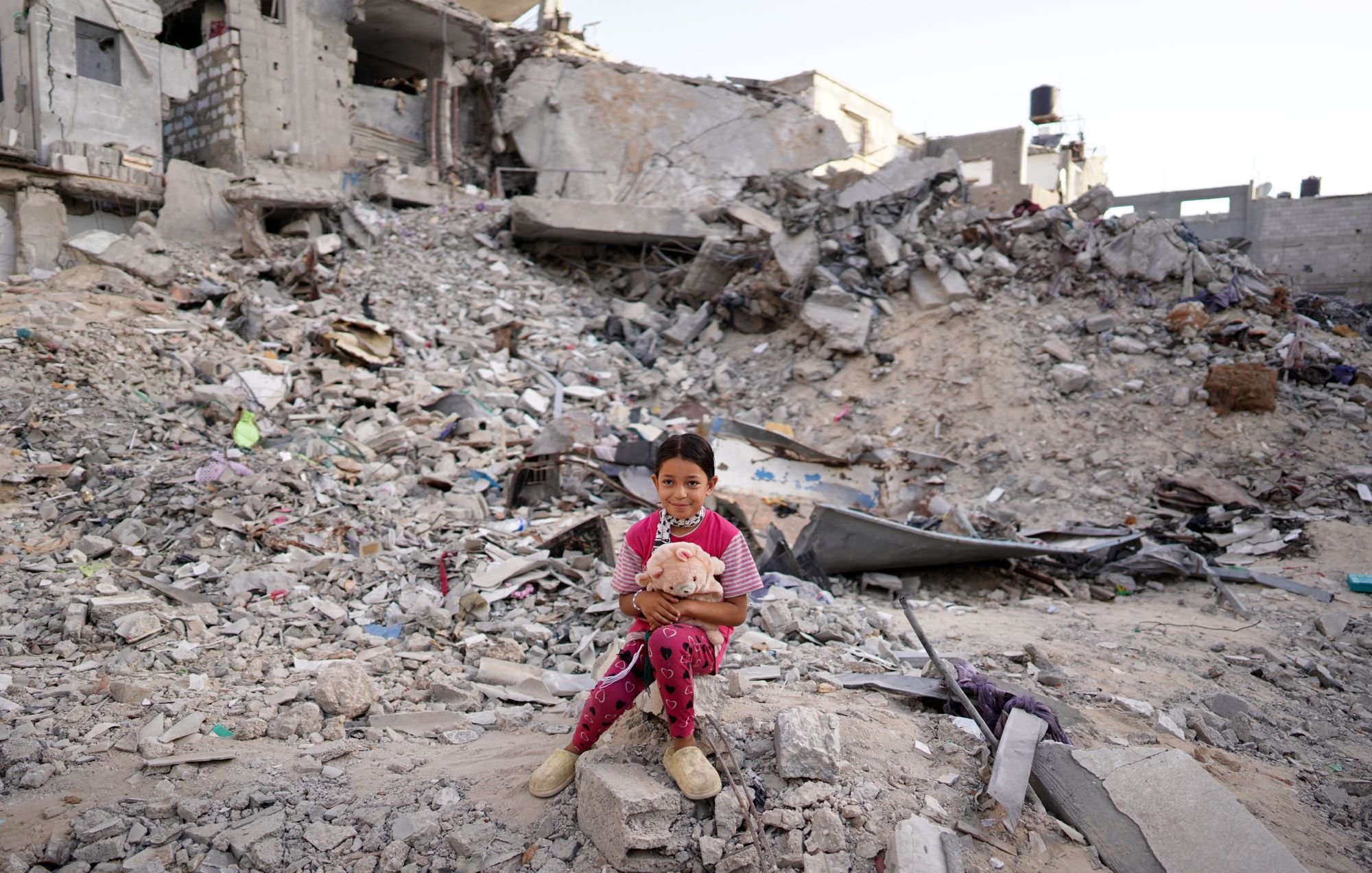 در این تصویر، دخترکی کوچک بر روی زمین در میان ویرانه‌های یک ساختمان نشسته است. در پس‌زمینه، تخریب‌های وسیع و انبوهی از آجرها و مصالح ساختمانی دیده می‌شود. دخترک با لباسی صورتی و شلوار آبی، عروسکی در آغوش دارد و به دوربین نگاه می‌کند. وضعیت اطراف او به وضوح نشان‌دهنده ویرانی و داغی ناشی از یک حادثه یا جنگ است. احساساتی از معصومیت و ناامیدی در این تصویر قابل مشاهده است.