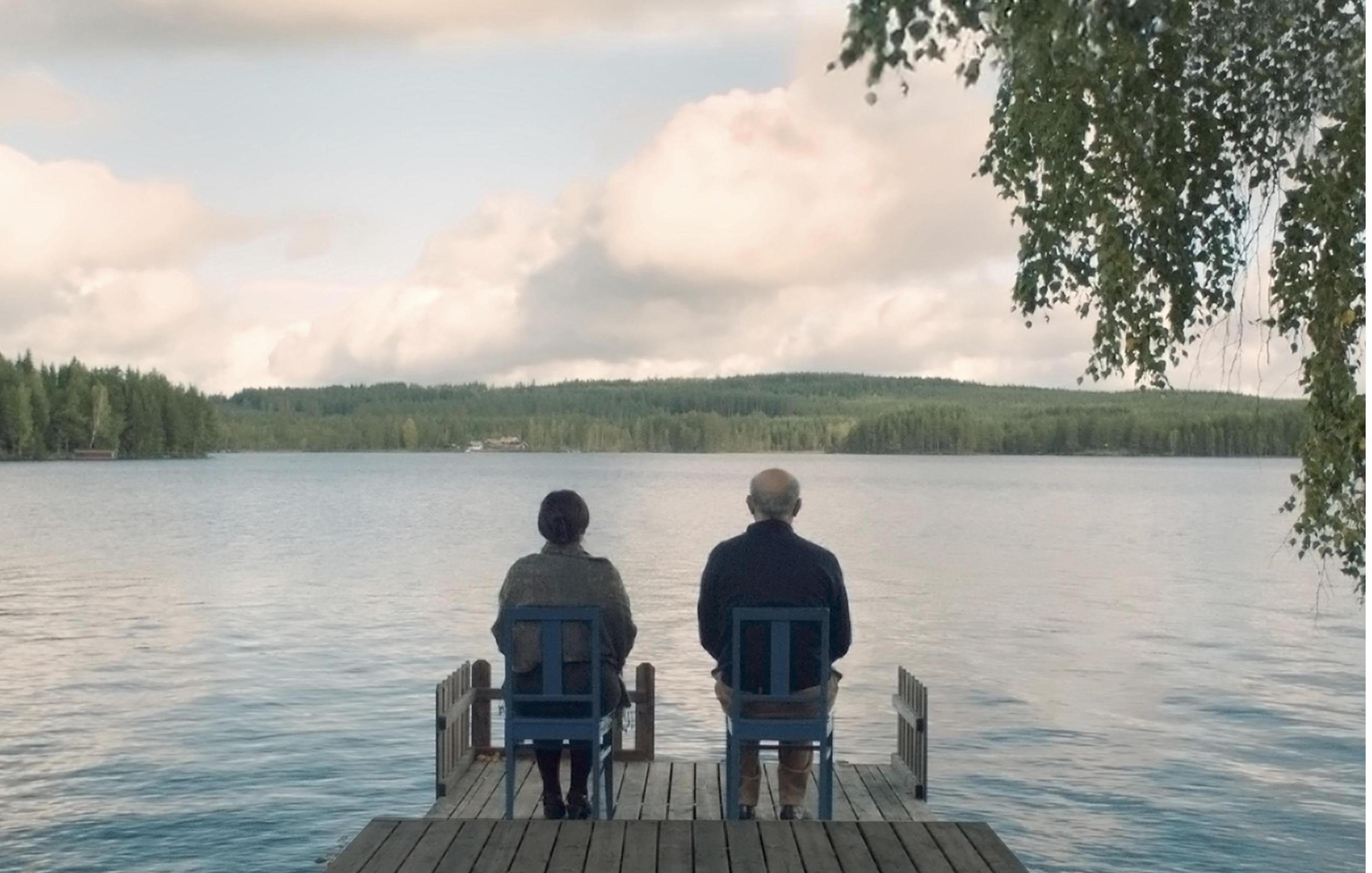 L'image montre deux personnes assises sur des chaises en bois sur une jetée qui s'avance dans un lac paisible. Ils regardent vers l'horizon, avec des arbres verdoyants en arrière-plan et un ciel nuageux au-dessus. L'atmosphère est calme et sereine, évoquant un moment de contemplation ou de discussion tranquille.