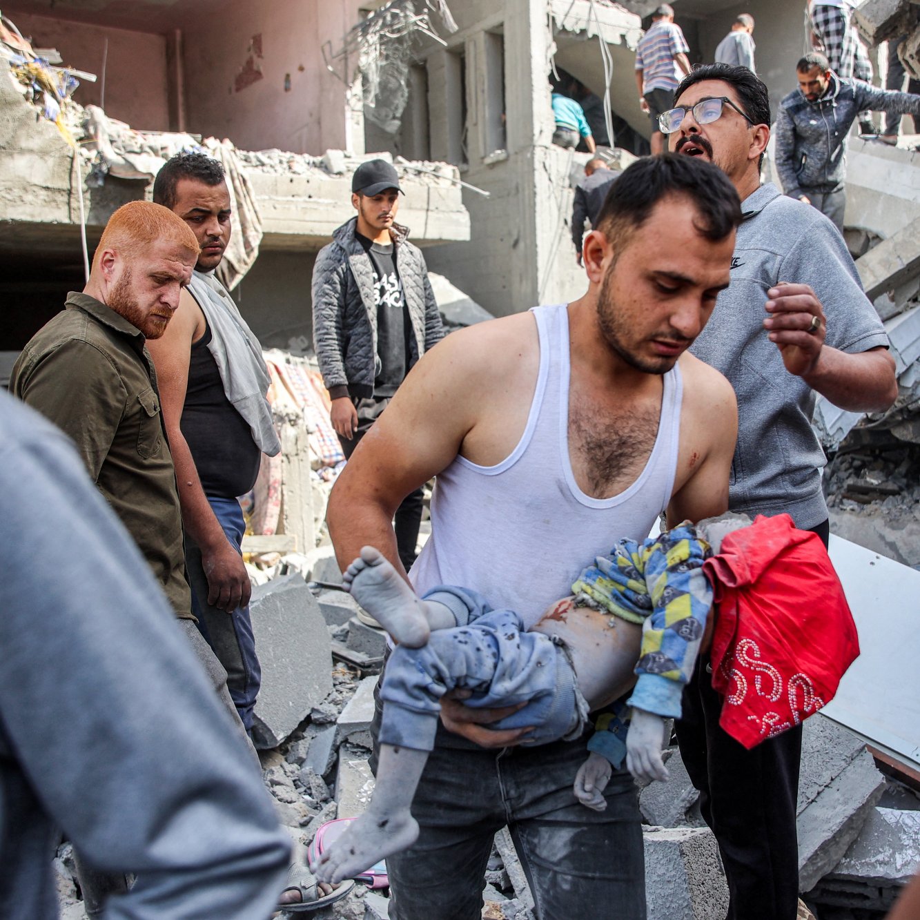 L'image montre une scène de désastre, probablement suite à un bombardement ou un tremblement de terre. On voit des décombres partout, avec des bâtiments endommagés. Un homme porte un enfant, probablement blessé, et semble préoccupé. D'autres personnes autour semblent s'affairer, cherchant à aider ou à évacuer les victimes. L'atmosphère est sombre et tragique, reflétant la détresse des personnes présentes.