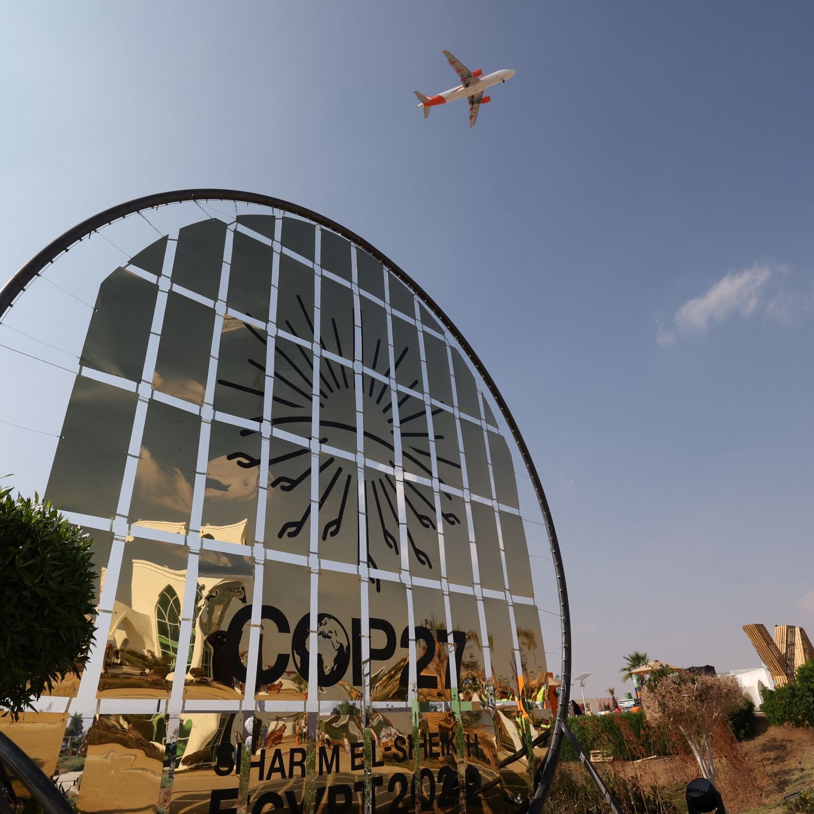 L'image montre un panneau de bienvenue à la COP27, ornée d'un design en miroir qui reflète le ciel. On aperçoit un avion volant au-dessus, suggérant une activité aérienne dans la région. Le paysage présente des éléments végétaux, comme des plantes et des arbres, et des personnes portant des vestes de sécurité sont visibles, indiquant une ambiance de préparation pour l'événement. Le ciel est clair avec quelques nuages.
