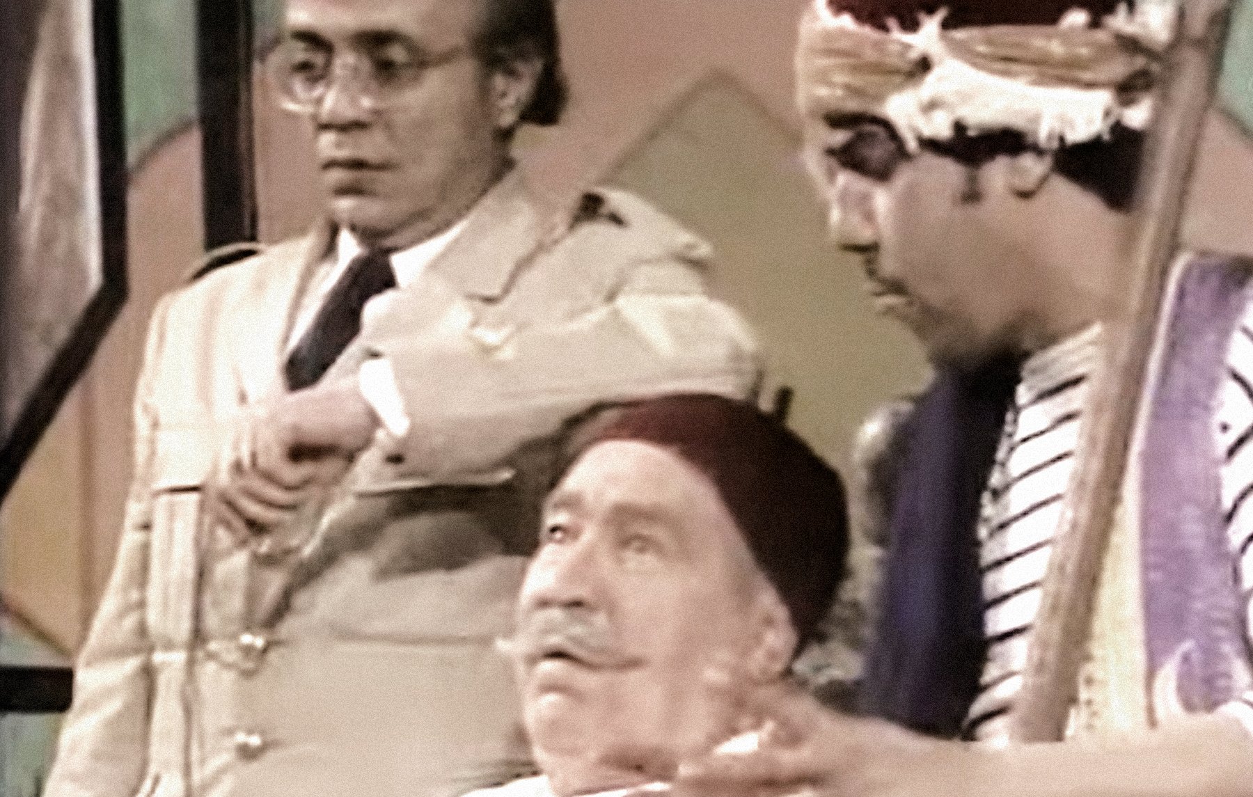 L'image montre trois personnages dans un cadre qui semble être une scène de théâtre ou une comédie. L'un des personnages est assis, portant une blouse blanche et un fez, affichant une expression détendue. À sa gauche se trouve un personnage en tenue militaire, regardant sa montre avec une expression préoccupée. À droite, un troisième personnage, également en fez, joue d'un instrument de musique tout en gesticulant, ajoutant une ambiance dynamique à la scène. L'ensemble évoque un moment comique ou dramatique.