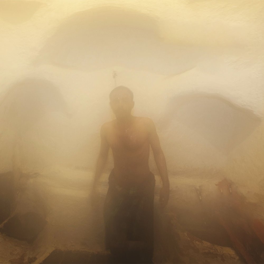 L'image montre un homme dans un environnement humide et brumeux, probablement à l'intérieur d'un hammam ou d'un sauna. La vapeur crée une atmosphère indistincte et floue, rendant les contours des objets difficilement visibles. L'homme est torse nu et semble se tenir debout, enveloppé par la vapeur qui l'entoure, ajoutant une sensation de chaleur et de sérénité à la scène. Les murs sont d'apparence texturée, typiques des espaces de bien-être.