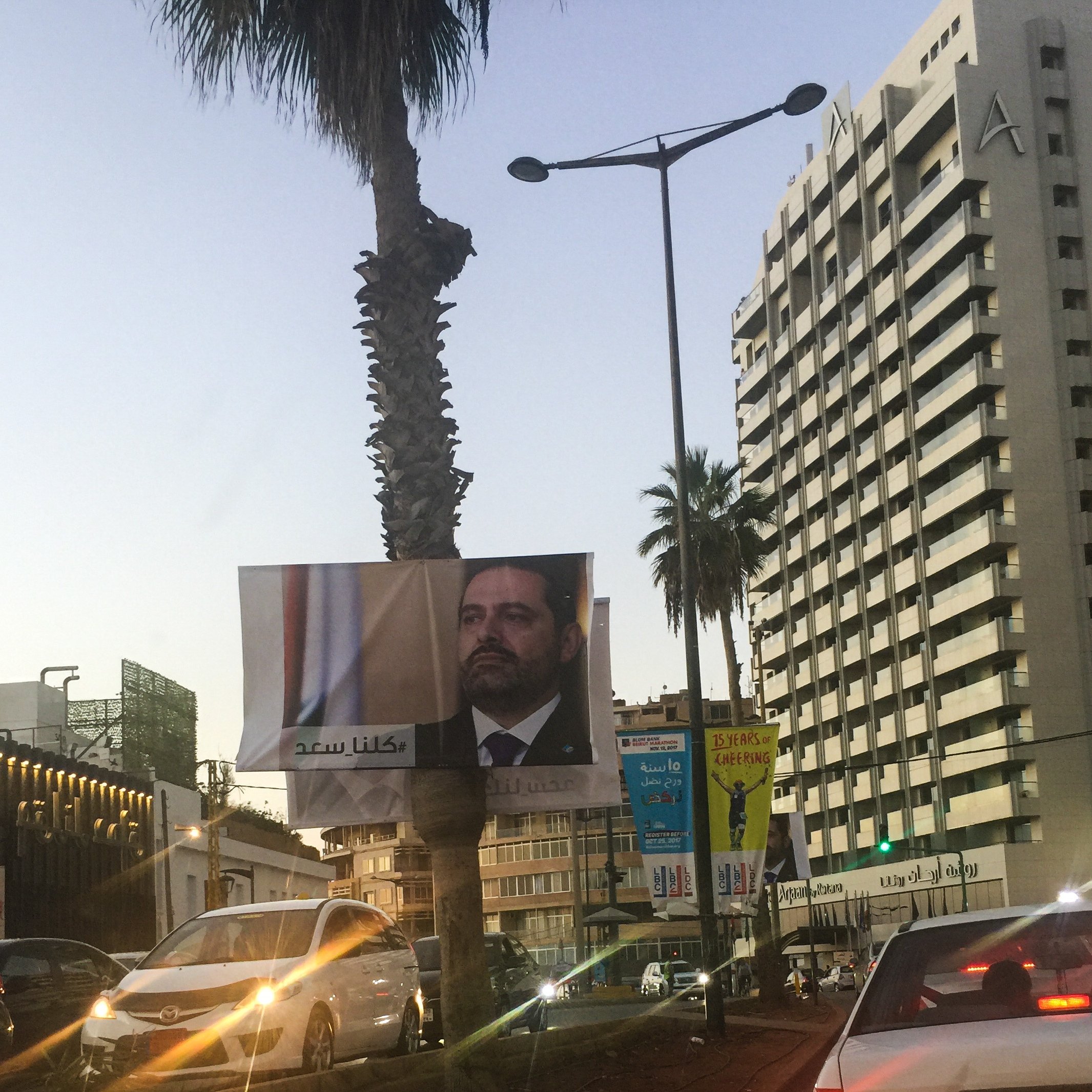 L'image montre une vue urbaine avec un grand panneau publicitaire affichant le visage d'un homme en costume, probablement un homme politique, et des arbres de palmiers sur le côté. En arrière-plan, on peut voir un immeuble de grande hauteur avec des enseignes, et d'autres panneaux d'affichage autour. La lumière du soir donne une ambiance douce à la scène. Les voitures sont visibles sur la route, suggérant une circulation urbaine.