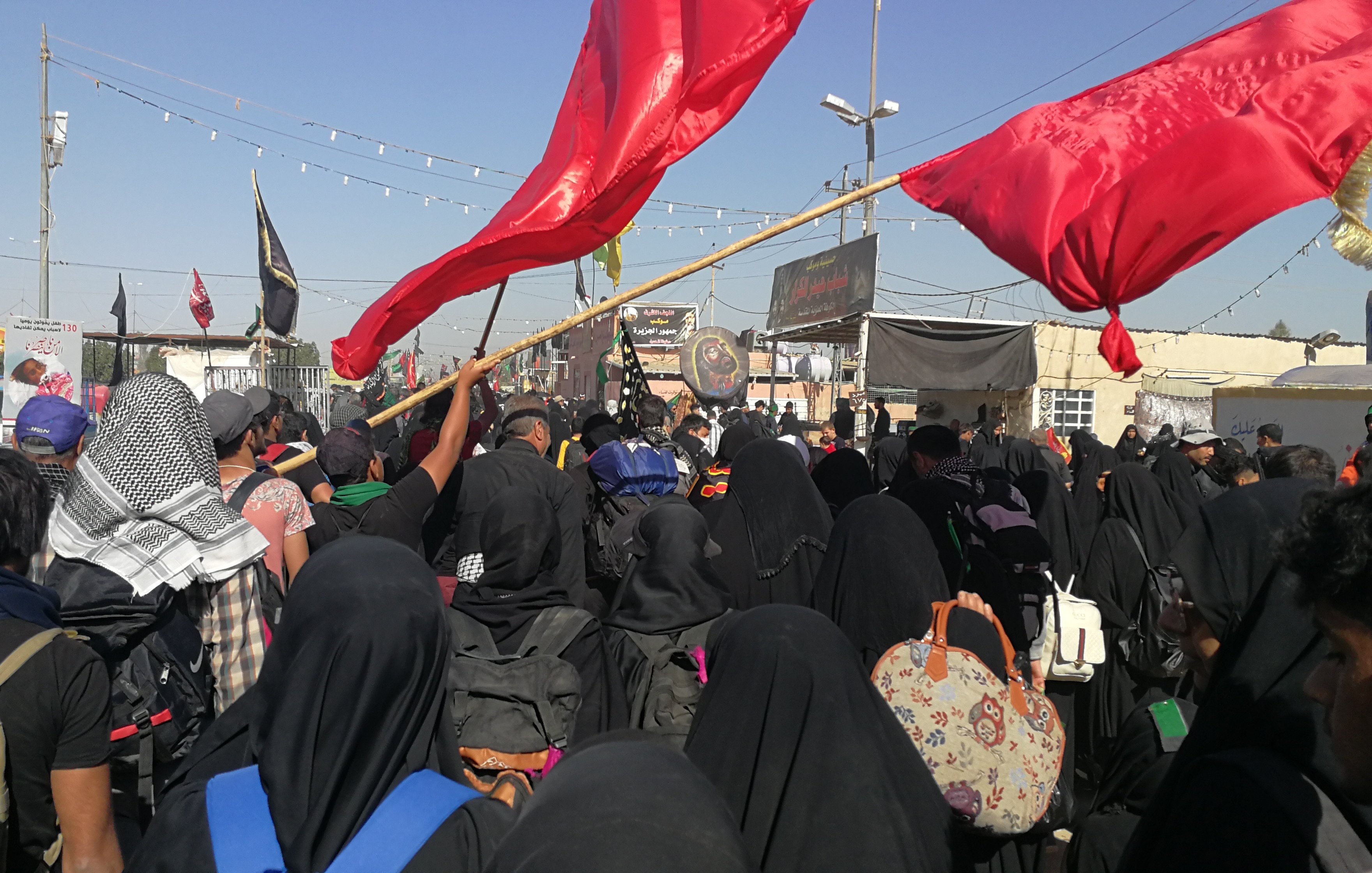 L'image montre une grande foule de personnes rassemblées, principalement vêtues de noir, ce qui suggère un événement commémoratif ou religieux. Plusieurs personnes tiennent des drapeaux rouges, et l'atmosphère semble être celle d'une manifestation ou d'un rassemblement significatif. Les participants portent des vêtements traditionnels et il y a des décors typiques de ce genre d'événement en arrière-plan, comme des bannières et éventuellement des symboles religieux. Le ciel est clair, ce qui indique une journée ensoleillée.