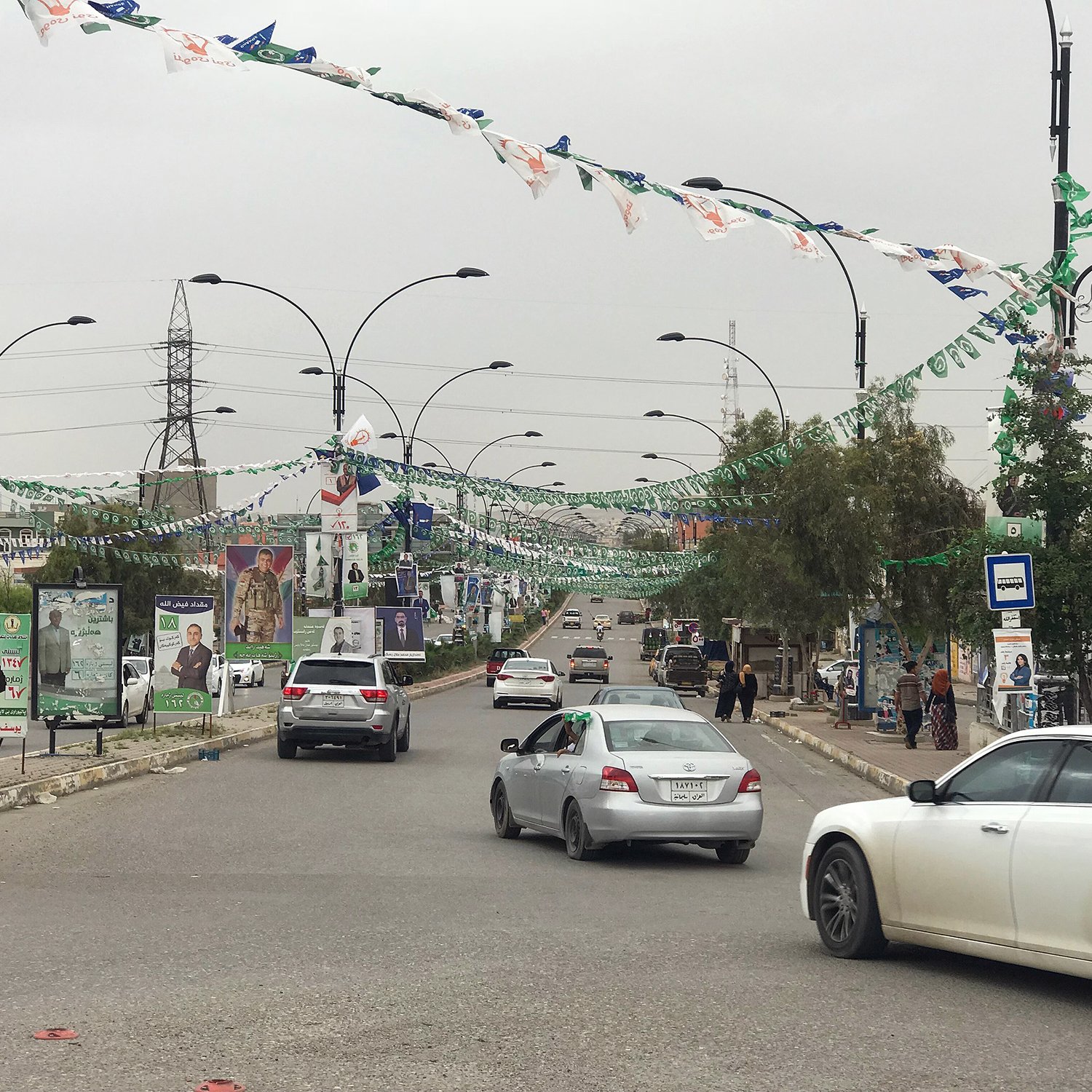 L'image montre une rue bordée de drapeaux et de bannières, probablement en préparation d'un événement ou d'une célébration. On peut voir plusieurs voitures circulant sur la route. À côté de la route, il y a des panneaux avec des affiches, ce qui indique une zone urbaine active. Des personnes se trouvent également sur le trottoir, et des arbres et des lampadaires sont présents le long de la rue, donnant un aspect dynamique à la scène. Le ciel semble nuageux, suggérant une ambiance calme.
