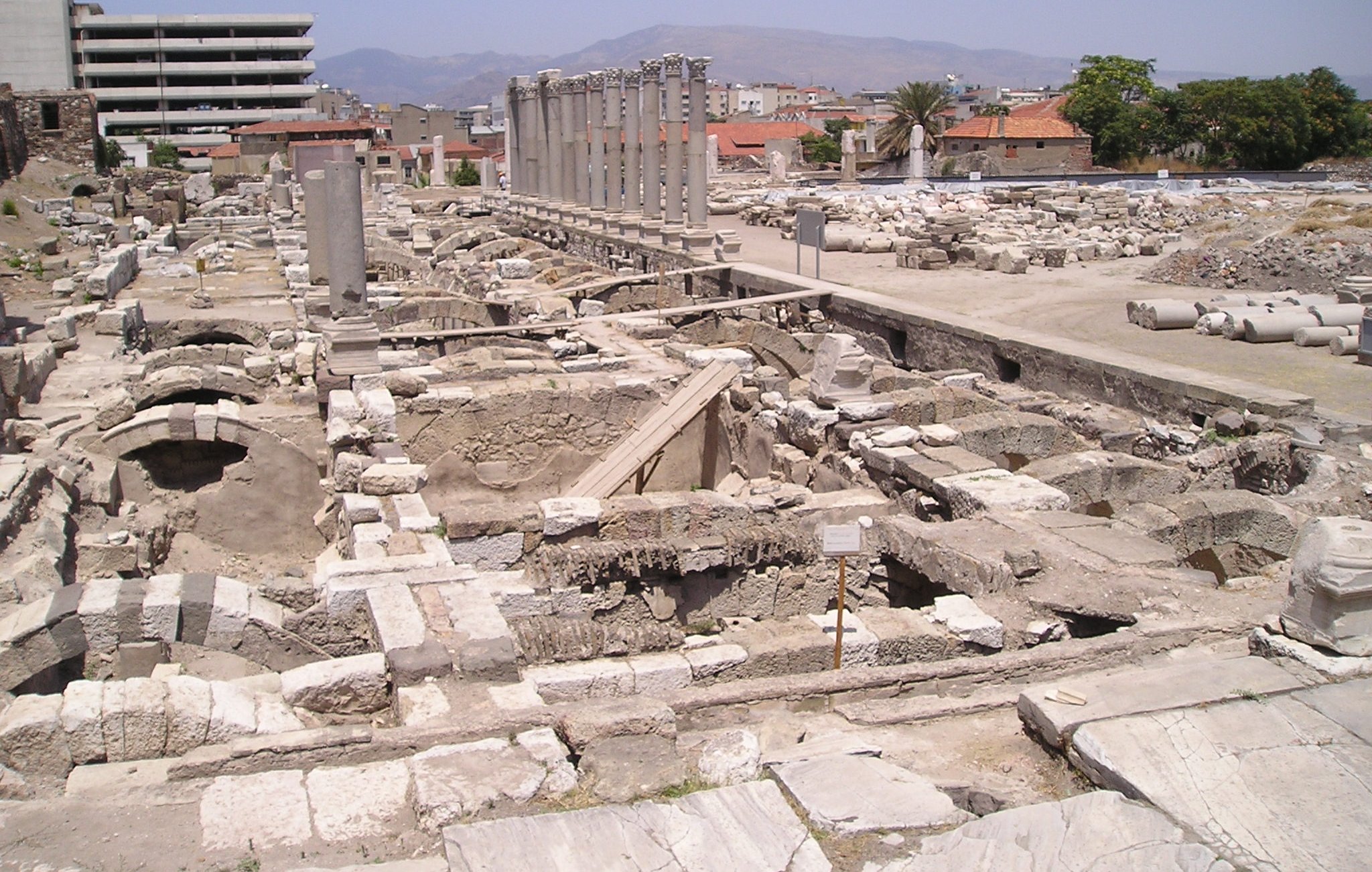 L'image montre un site archéologique avec des ruines et des structures anciennes. On aperçoit des colonnes dressées, probablement d'un bâtiment antique, ainsi que des fondations de murs en pierre. En arrière-plan, on peut distinguer des bâtiments modernes, contrastant avec les vestiges historiques. Le ciel est clair et le paysage est légèrement vallonné au loin. L'endroit semble être une zone de fouilles, avec des éléments qui témoignent d'une ancienne civilisation.