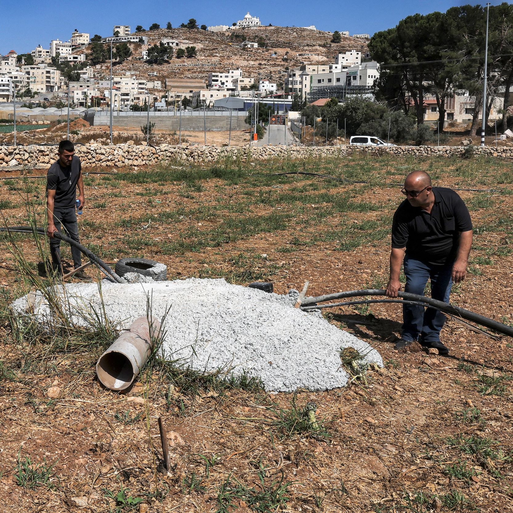 در این تصویر، دو مرد در حال کار بر روی یک زمین کشاورزی هستند. آنها مشغول بررسی یا نصب لوله‌هایی هستند که به یک سازه بتنی متصل شده است. درختان و گیاهان در اطراف وجود دارند و در پس‌زمینه، ساختمانی و تپه‌هایی دیده می‌شوند که حاکی از یک منطقه مسکونی است. آسمان آبی و صاف به نظر می‌رسد.