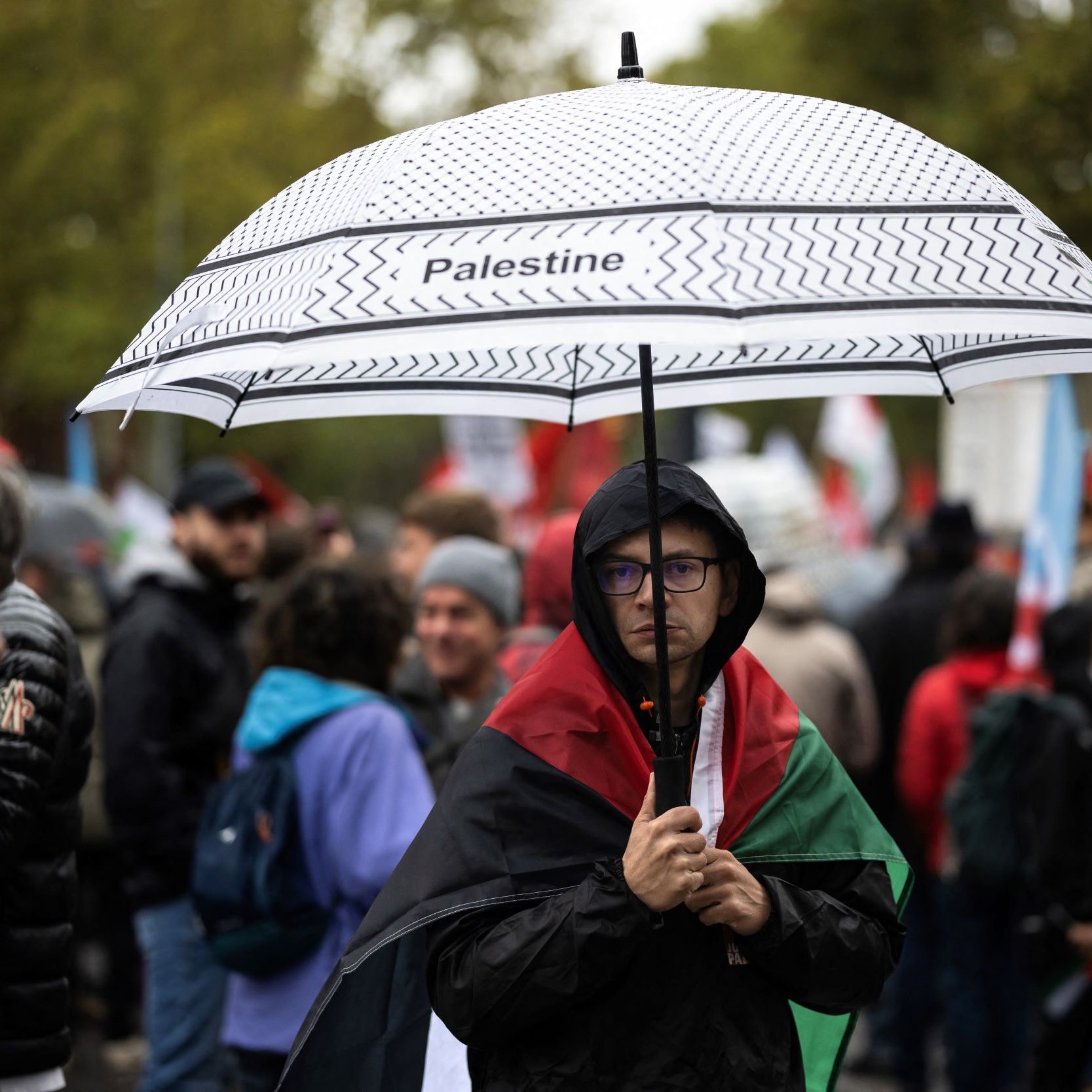 En la imagen, se puede ver a una persona que sostiene un paraguas con la palabra "Palestine" escrita en él. La persona lleva una bandera de Palestina sobre los hombros y parece estar en una manifestación o protesta. Alrededor se pueden observar otras personas, algunas con paraguas y banderas, en un ambiente cargado de emociones y solidaridad. El clima parece nublado o lluvioso, lo que añade a la atmósfera del evento.