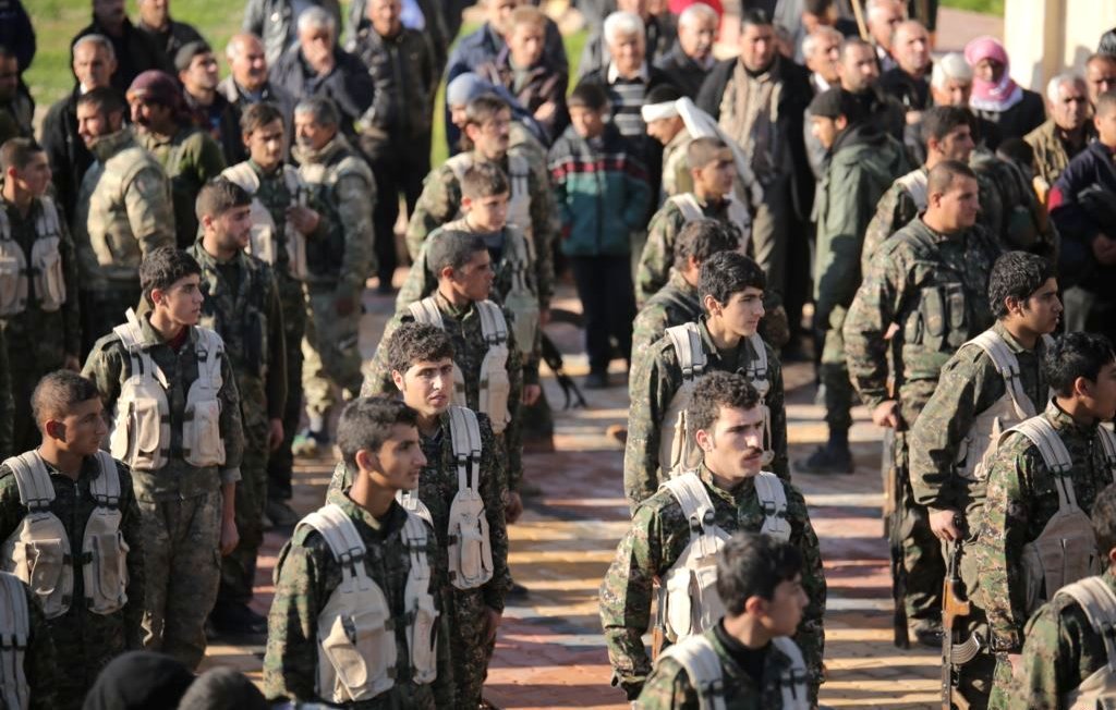 L'image montre un groupe de soldats en formation, portant des uniformes militaires et des équipements. Ils sont alignés, en regardant droit devant eux, et semblent être en train de participer à une cérémonie ou à un événement officiel. En arrière-plan, on peut observer une foule de personnes, probablement des civils, qui les regardent. L'atmosphère semble sérieuse et solennelle.