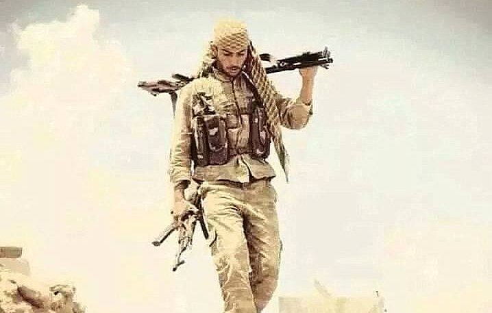 L'image montre un homme portant une tenue militaire, avec un chèche sur la tête. Il marche sur un terrain désertique, tenant deux armes, l'une à la main et l'autre portée sur son épaule. Son expression semble déterminée et il est entouré d'un paysage aride. L'atmosphère de l'image évoque un contexte de conflit ou de guerre.