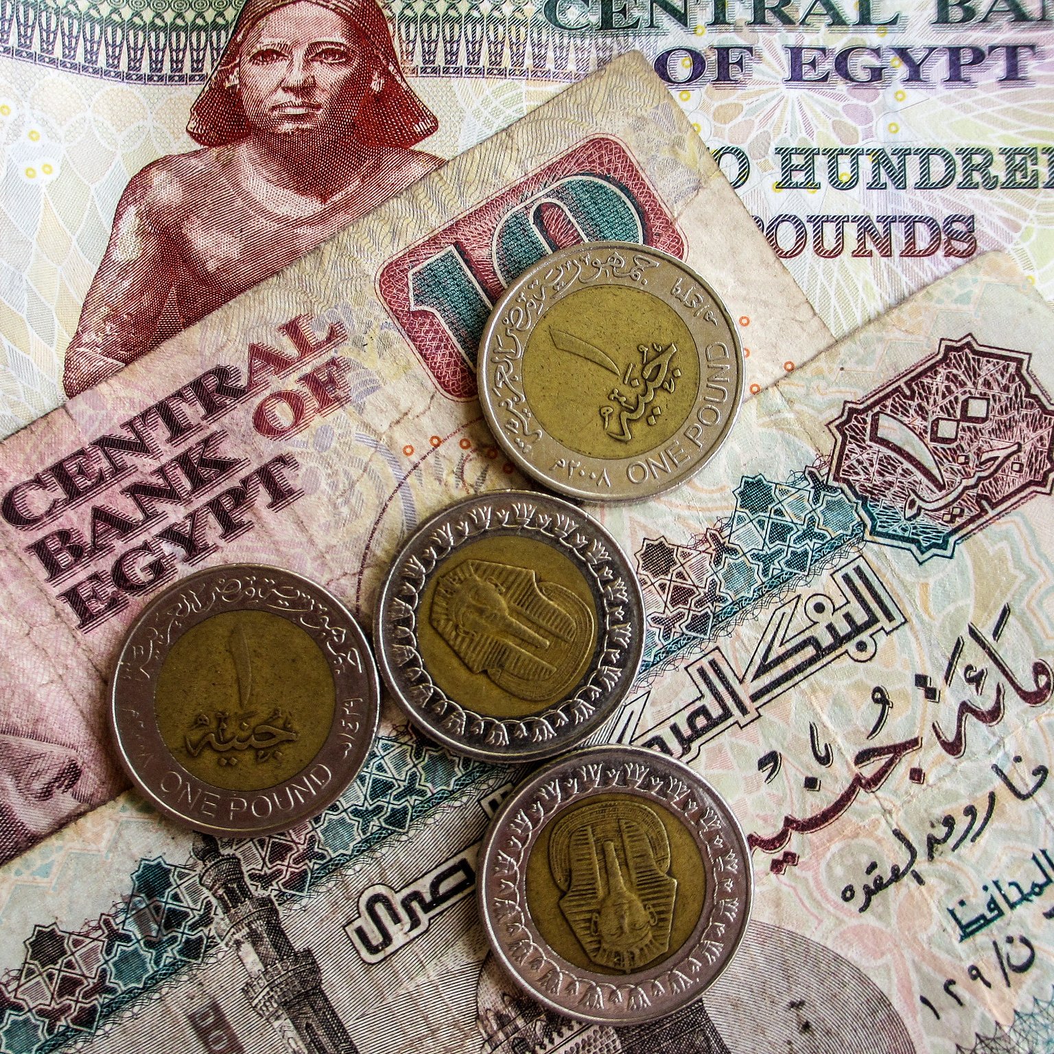 L'image montre des billets et des pièces de monnaie égyptiens. On peut voir un billet de 100 livres égyptiennes, ainsi qu'un billet de 10 livres. En dessous, il y a des pièces de monnaie d'un pound. Les billets sont ornés de motifs et de textes en arabe, indiquant qu'ils proviennent de la Banque Centrale d'Égypte. L'ensemble présente une variété de couleurs et de designs typiques de la monnaie égyptienne.