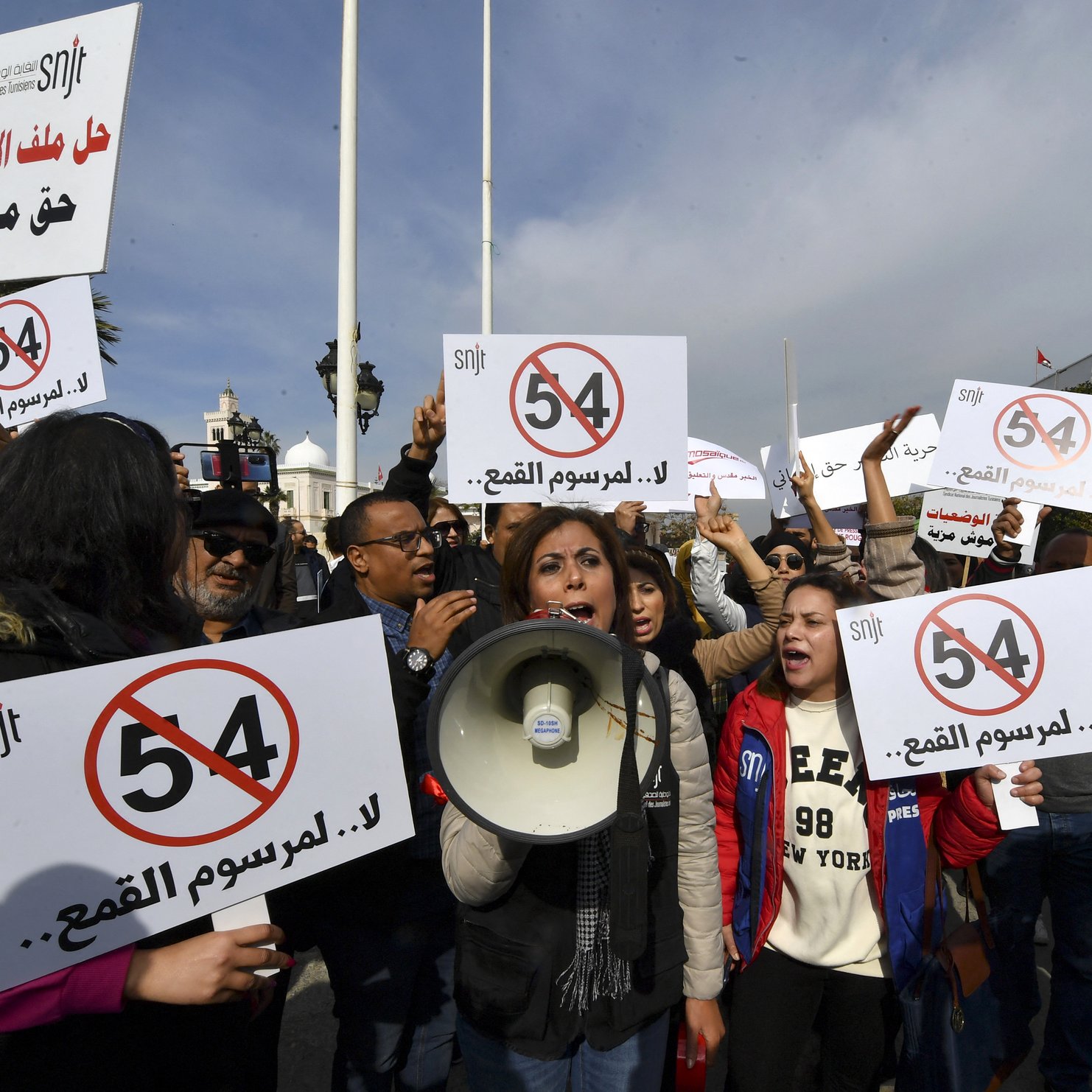 L'image montre une manifestation avec des groupes de personnes tenant des pancartes. Une femme au centre utilise un mégaphone pour s'exprimer. Les pancartes affichent des messages en arabe et en chiffres, notamment le chiffre "54", ainsi que des slogans contre "la censure". L'atmosphère semble énergique, avec des participants engagés dans la cause. Les drapeaux au fond ajoutent à l'ambiance de protestation.