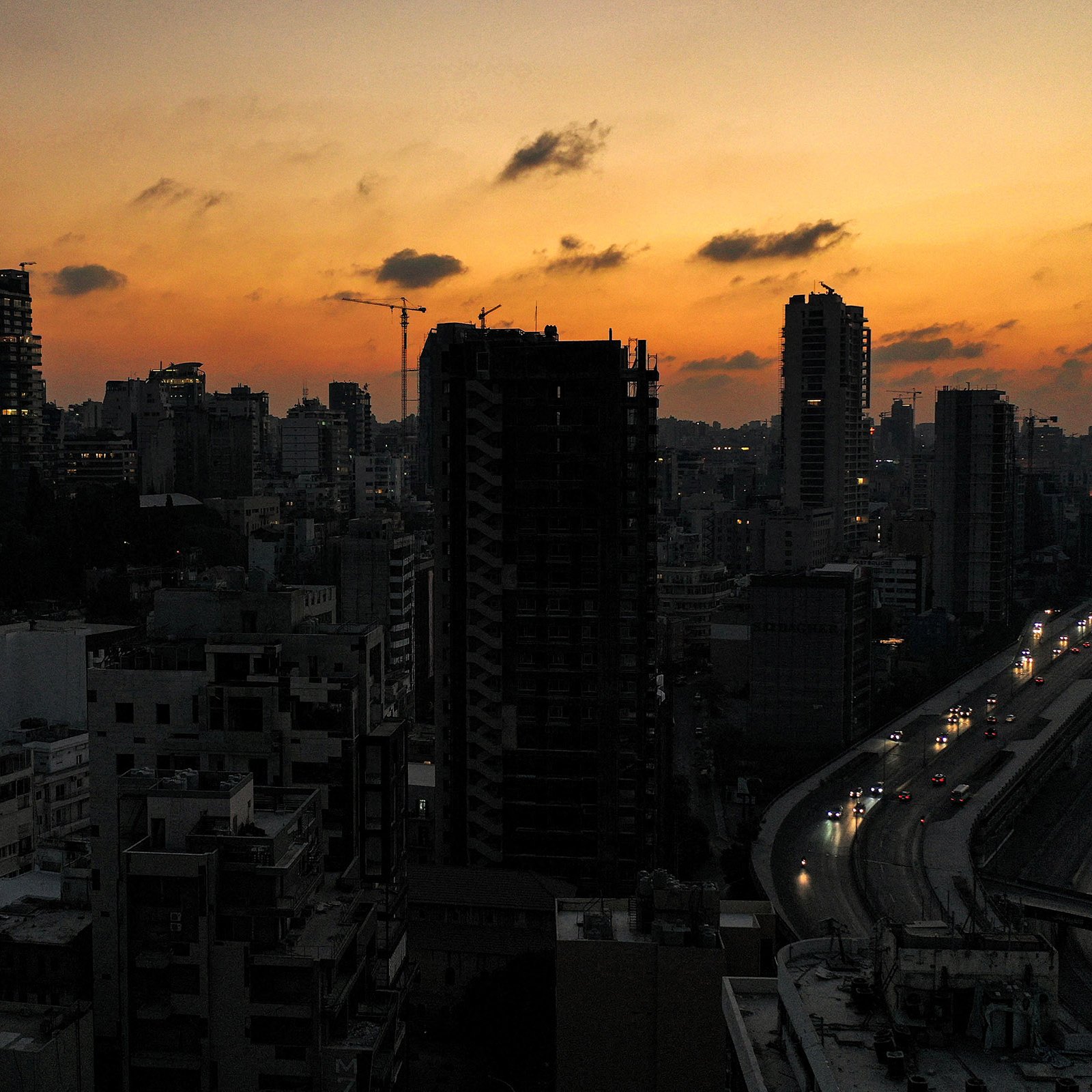 تظهر الصورة مشهداً ليلياً لمدينة مكتظة بالمباني والعمارات. السماء تتلون بألوان الغروب، حيث تتدرج الألوان من البرتقالي إلى الأزرق الداكن. في الأسفل، يمكن رؤية طريق يمر بجانب المباني، وقد ظهرت بعض السيارات وهي تسير في الاتجاهات المختلفة. الأضواء بدأت تتلألأ في النوافذ، مما يضفي على المشهد جوًا ساحرًا يعكس حيوية المدينة في المساء.