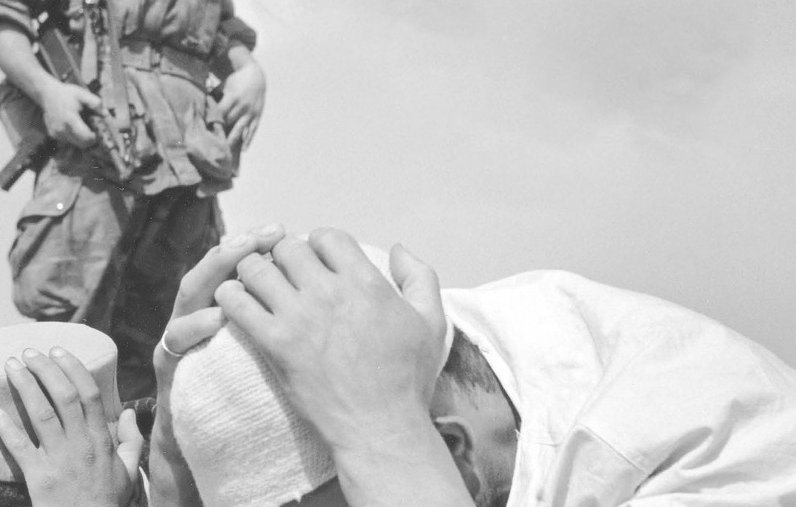 L'image montre un soldat en uniforme tenant une arme, se tenant en arrière-plan, tandis que deux personnes au premier plan sont inclinées vers le sol. L'une d'elles semble tenir sa tête dans ses mains, exprimant une grande détresse ou un sentiment de soumission. L'ambiance générale semble chargée d'émotion et de tension, suggérant une situation de conflit ou de crise. Les éléments monochromes de la photographie renforcent l'intensité de la scène.