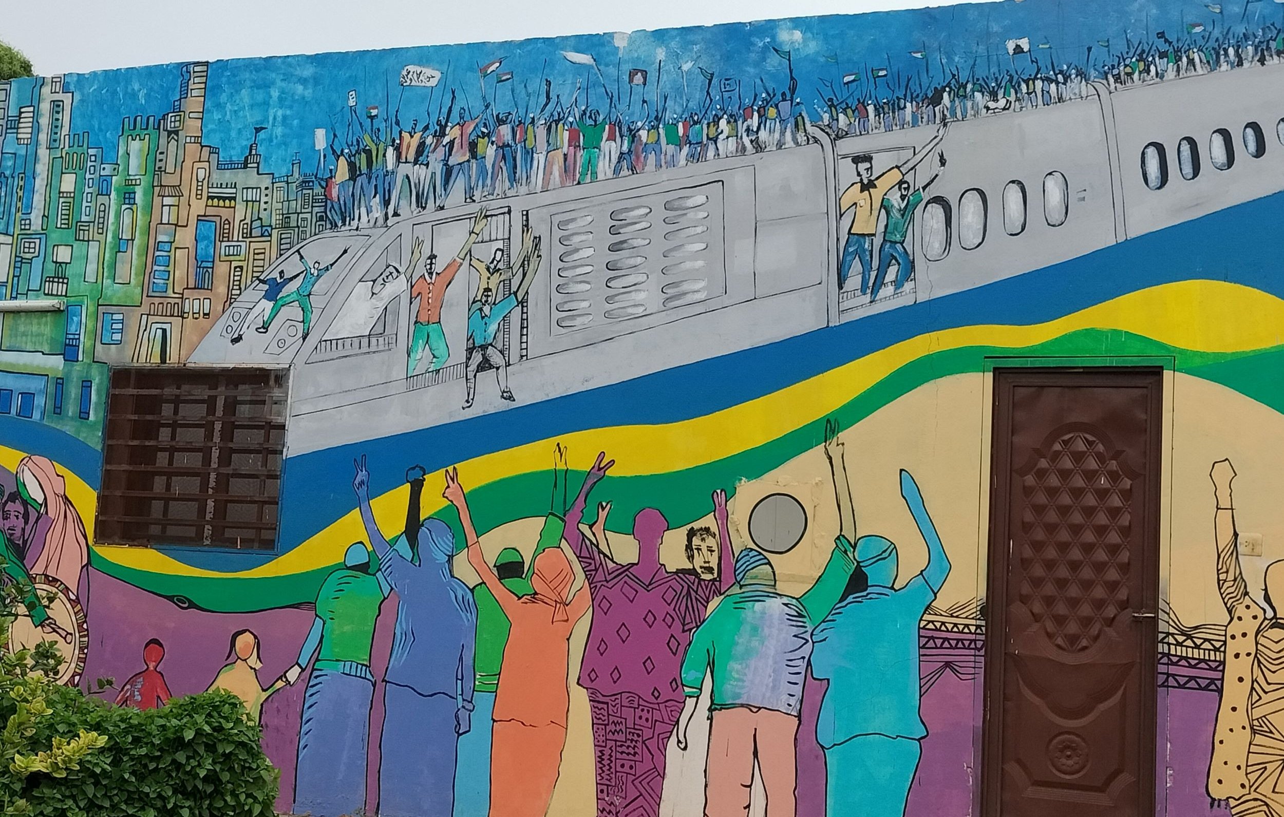 L'image montre une fresque murale colorée qui illustre une scène dynamique. On y voit des personnes célébrant et manifestant, les bras levés, sur un fond représentant un avion et une ville. Les couleurs vives ajoutent une énergie palpable à la scène, reflétant un sentiment de joie ou de solidarité. Des silhouettes de différents personnages sont visibles, et l'ensemble de l'œuvre évoque un message de rassemblement et d'espoir.