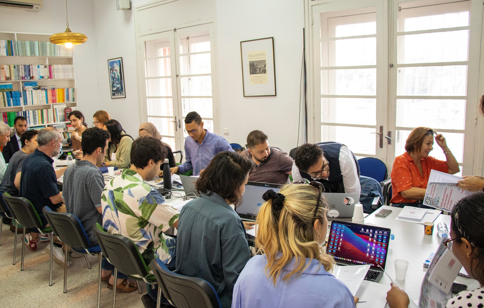 L'image montre une salle de réunion où un groupe de personnes est assis autour d'une grande table. Plusieurs participants semblent travailler sur des ordinateurs portables, tandis que d'autres consultent des documents. Les murs sont décorés de livres et d'illustrations, et plusieurs fenêtres laissent entrer la lumière naturelle. L'ambiance apparaît studieuse et collaborative.