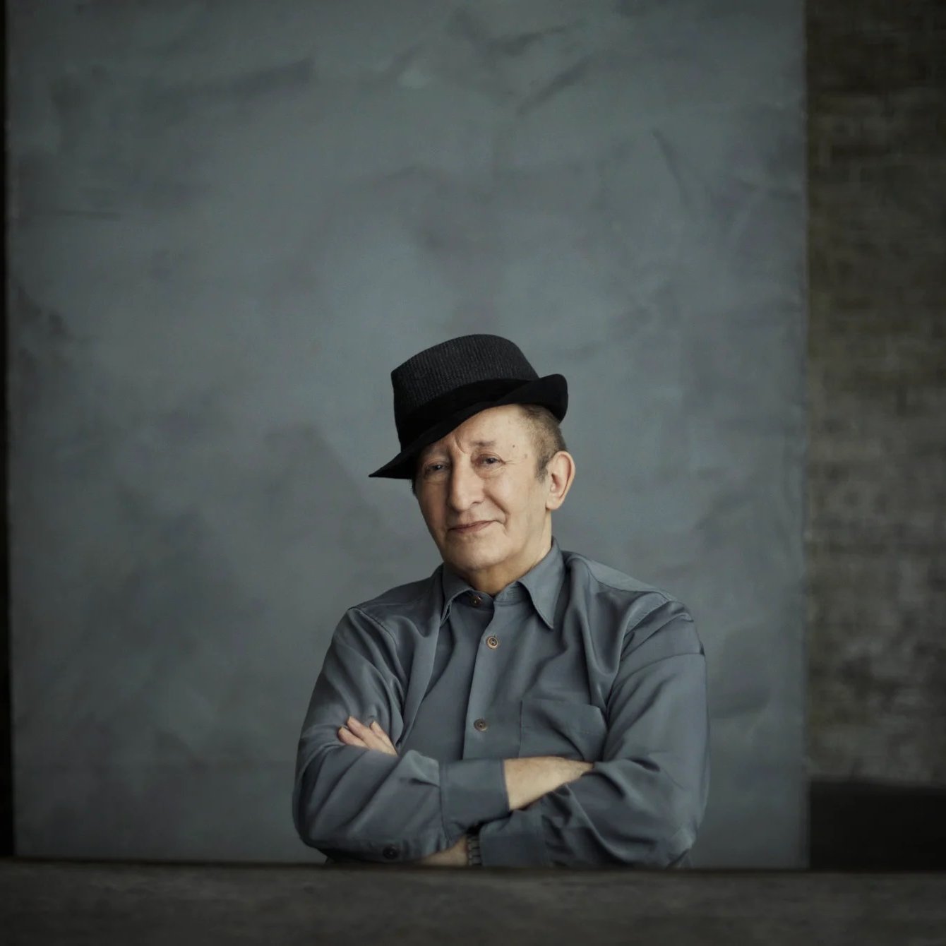 L'image montre un homme assis avec un chapeau noir. Il a un léger sourire et les bras croisés sur une table. Il porte une chemise grise. L'arrière-plan est simple, avec une texture de mur gris qui crée une atmosphère sobre et contemplative. La lumière est douce, accentuant les traits de son visage.