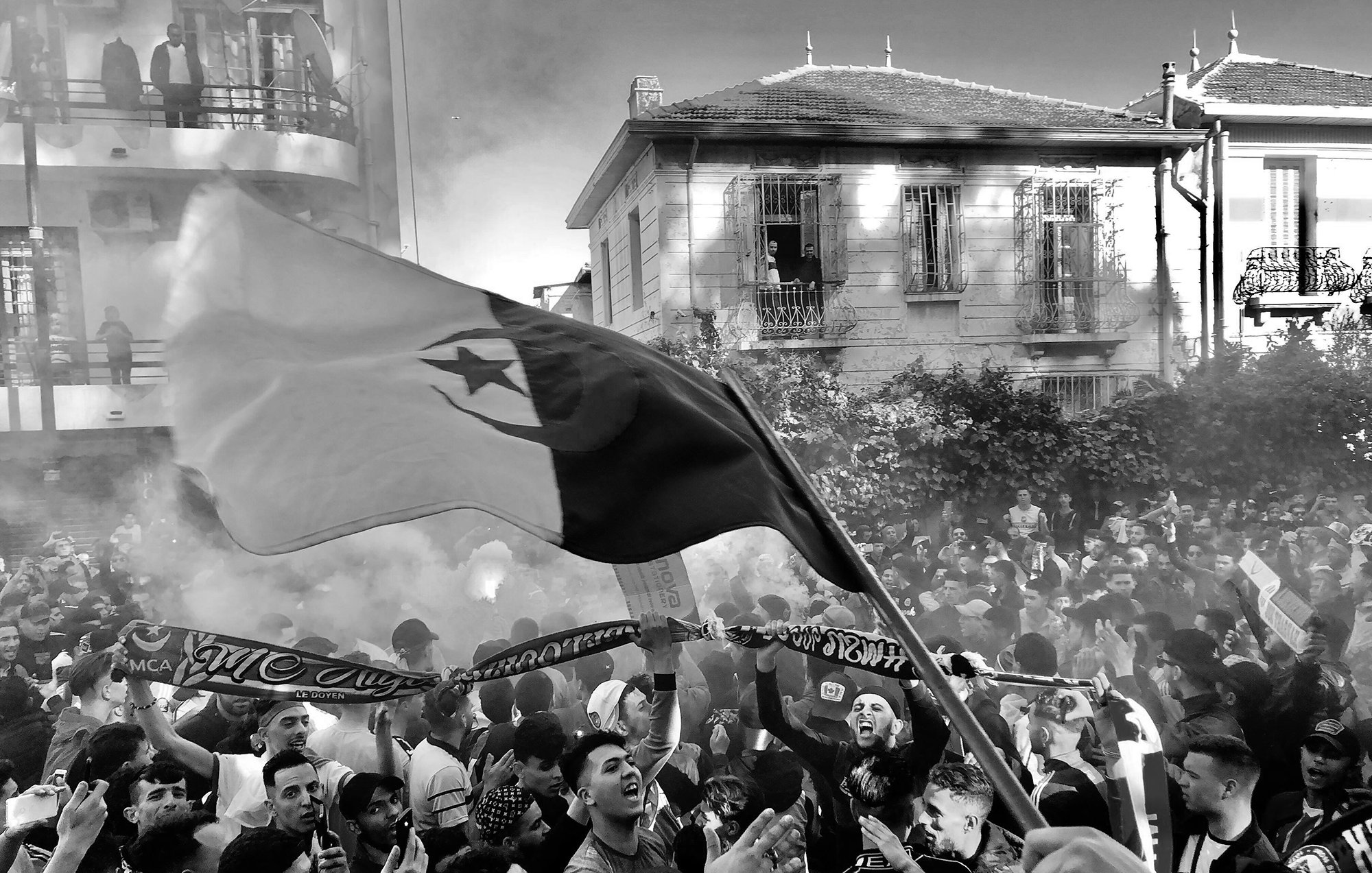 تظهر في الصورة حشود من الناس يحتفلون، مع مزيج من الدخان في الهواء. تتواجد مجموعة من المشجعين يحملون الأعلام واللافتات. يبدو أن هناك أجواء حماسية، مع تفاعل كبير بين المشاركين في الاحتفال. الخلفية تضم مباني سكنية، مما يضيف طابعاً حضرياً إلى المشهد.