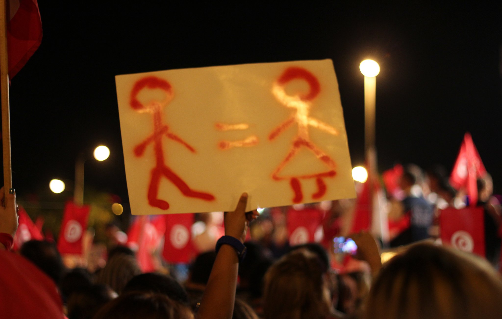 L'image montre une manifestation nocturne avec des gens rassemblés, portant des drapeaux rouges. Au premier plan, une personne tient une pancarte sur laquelle sont dessinées deux silhouettes : un homme et une femme, accompagnés du symbole "=" entre eux. Cela semble représenter un message d'égalité entre les sexes. L'ambiance est animée, et le public semble engagé sur le sujet.
