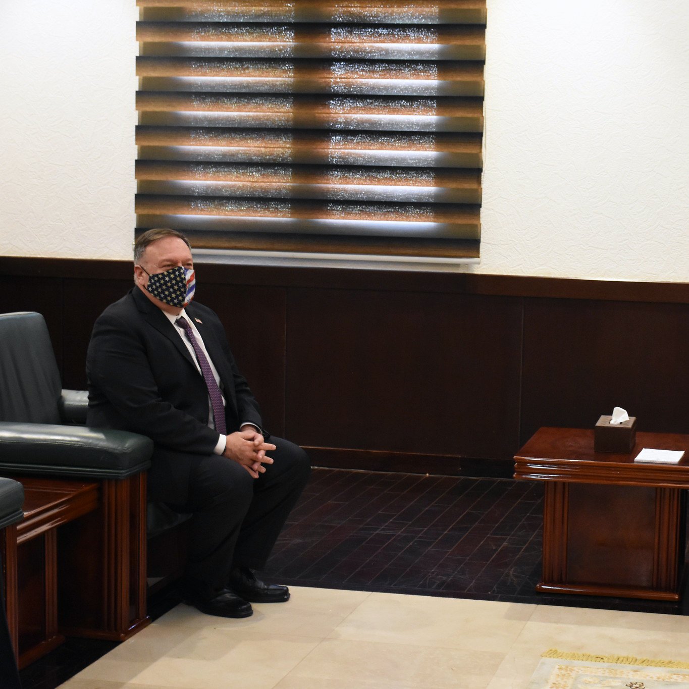 در این تصویر، دو مرد در یک اتاق رسمی نشسته‌اند. یکی از آنها که سمت چپ تصویر است، ماسک دارد و ظاهری رسمی با کت و شلوار پوشیده است. مرد دیگر در سمت راست نیز لباس رسمی به تن دارد و به طرف مرد اول نگاه می‌کند. در پس‌زمینه، پرده‌های پنجره و پرچم یک کشور مشخص است. فضا به وضوح رسمی و موقر به نظر می‌رسد و به احتمال زیاد مربوط به یک ملاقات دیپلماتیک است.