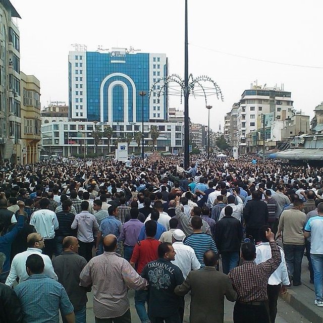 L'image montre une grande foule de personnes rassemblées dans une zone urbaine. On peut voir des bâtiments modernes en arrière-plan, ainsi qu'un aménagement urbain typique avec des lampadaires. La multitude semble engagée dans une manifestation ou un rassemblement, avec des gens de différentes orientations et tenues. L'ambiance générale évoque un moment de solidarité ou de protestation.