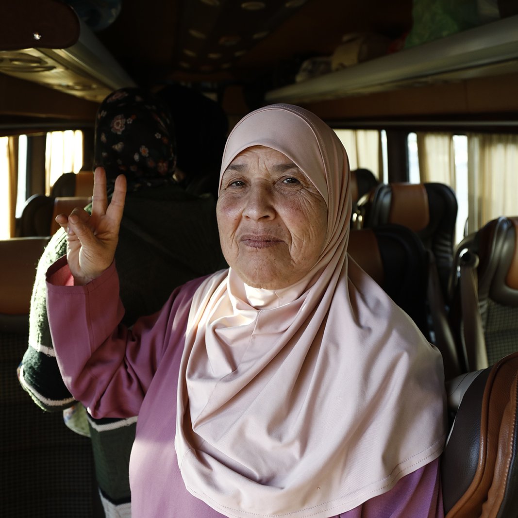 L'image présente une femme âgée à l'intérieur d'un bus. Elle porte un hijab de couleur claire et un vêtement de style traditionnel. Elle fait un signe de paix avec ses doigts tout en souriant. En arrière-plan, on peut apercevoir d'autres passagers et l'intérieur du bus avec des sièges confortables. L'atmosphère semble chaleureuse et amicale.