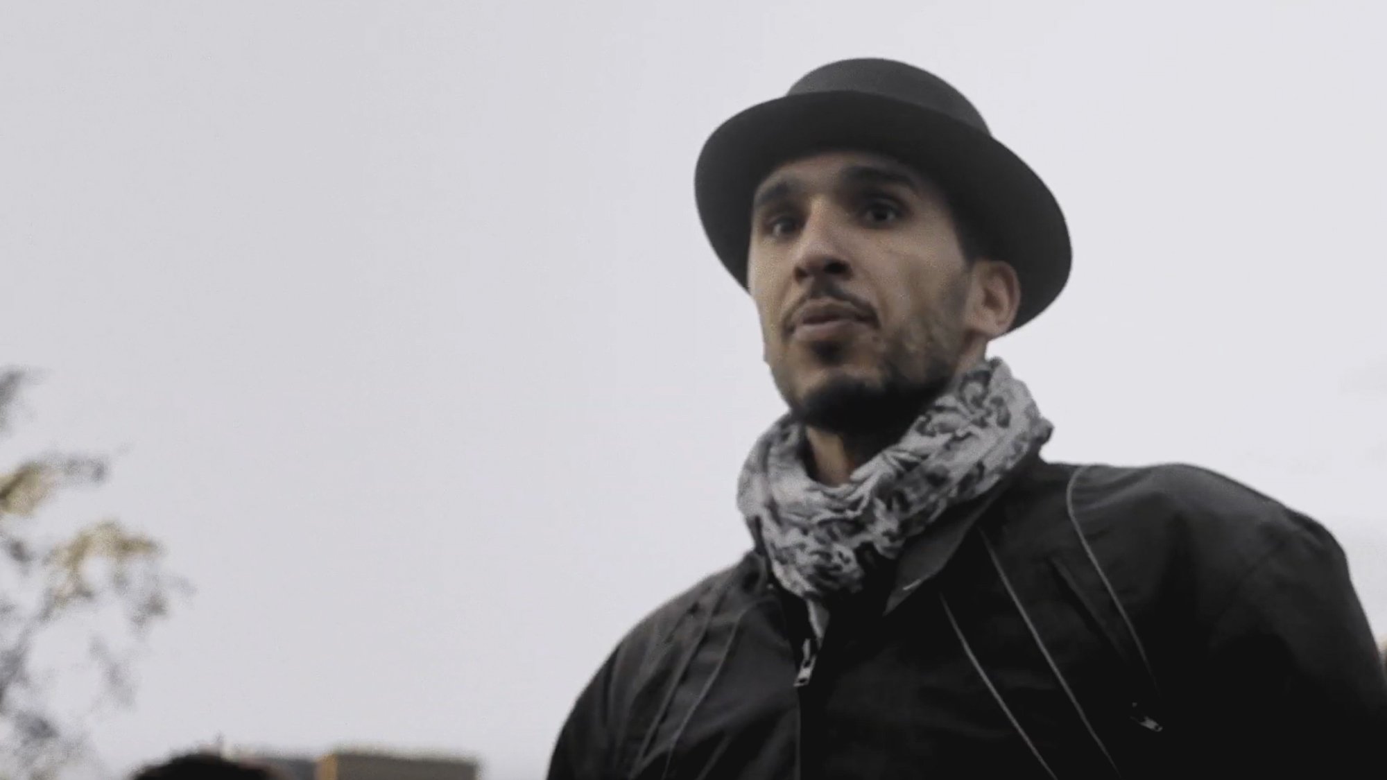 L'image montre un homme portant un chapeau noir et une écharpe. Il a une expression sérieuse et regarde droit devant lui. Le fond semble flou, suggérant une ambiance extérieure peut-être en milieu urbain ou dans un parc. Le temps semble nuageux, ce qui ajoute une atmosphère un peu sombre à la scène.