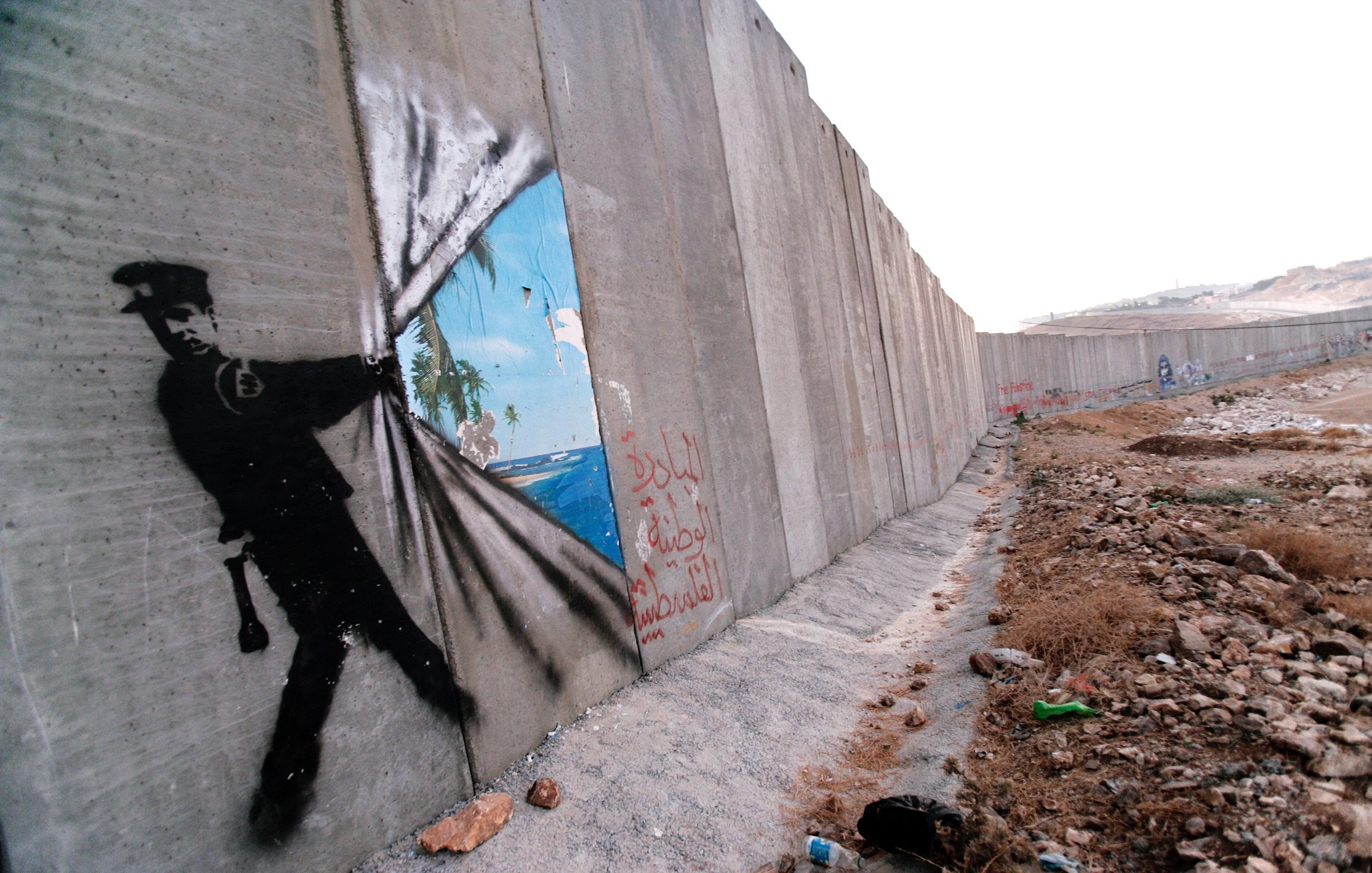 L'image montre un mur de béton, probablement un mur de séparation ou de sécurité, sur lequel est peint un graffiti représentant un policier. Ce dernier semble tirer un rideau pour révéler un paysage idyllique, avec des palmiers et un ciel bleu, symbolisant un contraste entre la réalité de l'environnement urbain et un espace rêvé ou libre. Sur le mur, on peut également voir des inscriptions en arabe. L'ensemble de l'image peut évoquer des thèmes de restriction, d'espoir et de liberté.