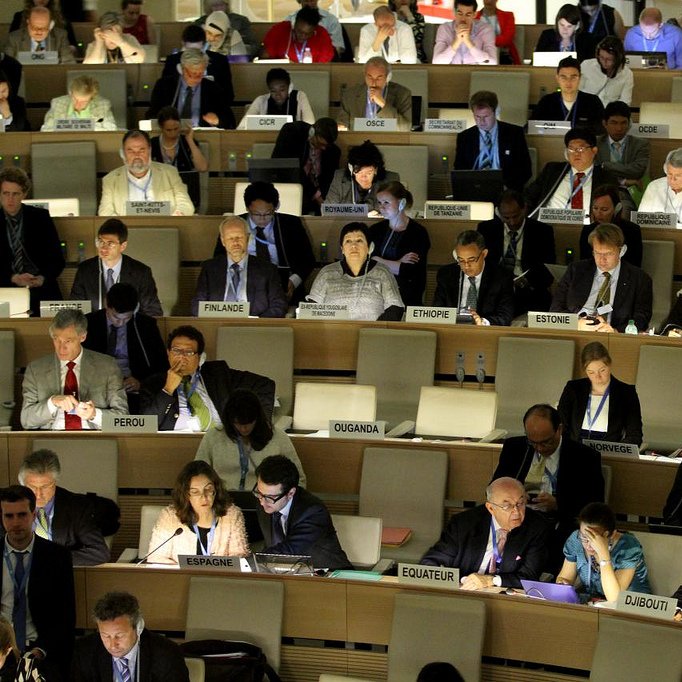 L'image montre une grande salle de conférence remplie de personnes assises à des tables. Les participants semblent concentrés sur leurs ordinateurs portables et divers documents, suggérant qu'ils sont engagés dans une discussion ou une réunion importante. À l'arrière-plan, on peut voir des drapeaux de différents pays visibles, ce qui indique un contexte international. L'éclairage de la salle est tamisé, créant une atmosphère sérieuse et studieuse.