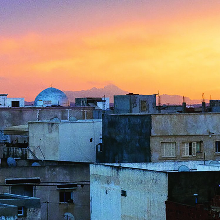 L'image montre un paysage urbain au coucher du soleil. On y voit plusieurs bâtiments aux façades de couleur beige et grise, avec des toits plats. À l'horizon, une belle teinte orange et rose illumine le ciel, créant une ambiance chaleureuse. Un dôme se distingue parmi les structures, suggérant la présence d'une mosquée ou d'un autre bâtiment significatif. Les lignes des constructions et le ciel coloré contrastent magnifiquement, donnant une impression de tranquillité.