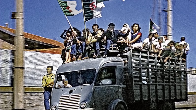 L'image montre un camion dont le plateau est rempli de personnes célébrant avec des drapeaux algériens. Les gens semblent joyeux et enthousiastes, tenant les drapeaux en l'air. En arrière-plan, on aperçoit des bâtiments et des pylônes électriques. La scène évoque une ambiance festive et patriotique. Les couleurs sont vives, et l'ensemble transmet un sentiment d'unité et de célébration.