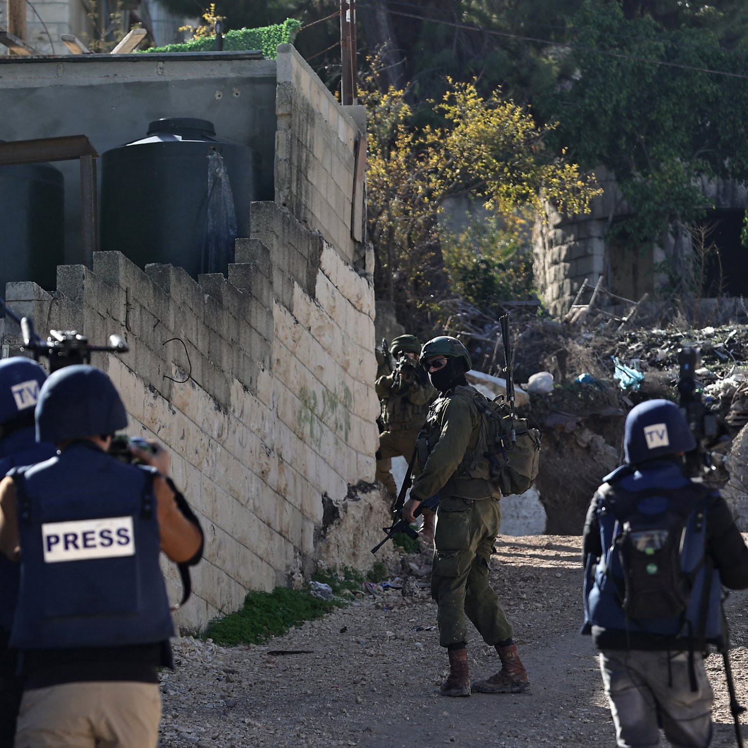 L'image montre une scène de conflit avec des soldats armés et des journalistes. Il y a des soldats en uniforme, portant des casques et des équipements de protection, qui se déplacent dans un environnement urbain, probablement dans une zone de conflit. Des journalistes sont également présents, identifiables grâce à leurs gilets marqués du mot "PRESS". En arrière-plan, on peut apercevoir des structures en ruine et des éléments de débris, ce qui suggère un contexte de tensions ou de violence.
