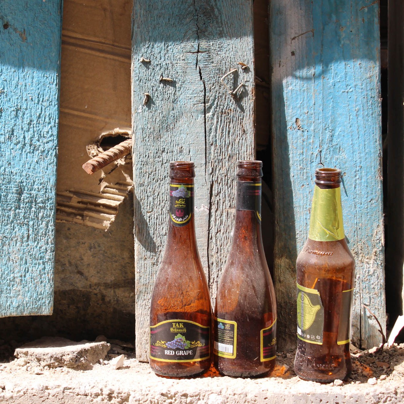 The image features three glass bottles positioned in front of a weathered blue wooden wall. The bottles appear dusty and have some remnants of liquid inside them. The wall shows signs of decay, with peeling paint and a small gap revealing damaged wood behind it. The ground is uneven, with dirt and debris scattered around, adding to the overall rustic and neglected feel of the scene.