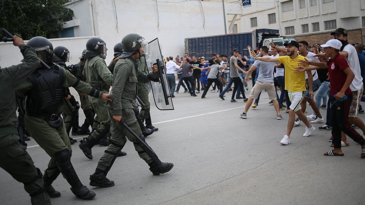 L'image montre une scène de confrontation entre des agents de sécurité en uniforme, portant des casques et des protections, et un groupe de manifestants. Les policiers avancent avec des boucliers, tandis que des personnes dans le public semblent manifester avec des gestes de défi. L'atmosphère est tendue, et le cadre urbain suggère un rassemblement intense, possiblement dans un contexte de protestation ou de manifestation. Les participants sont habillés de manière casual, et on peut voir une diversité dans leurs attitudes et leurs réactions face à la situation.