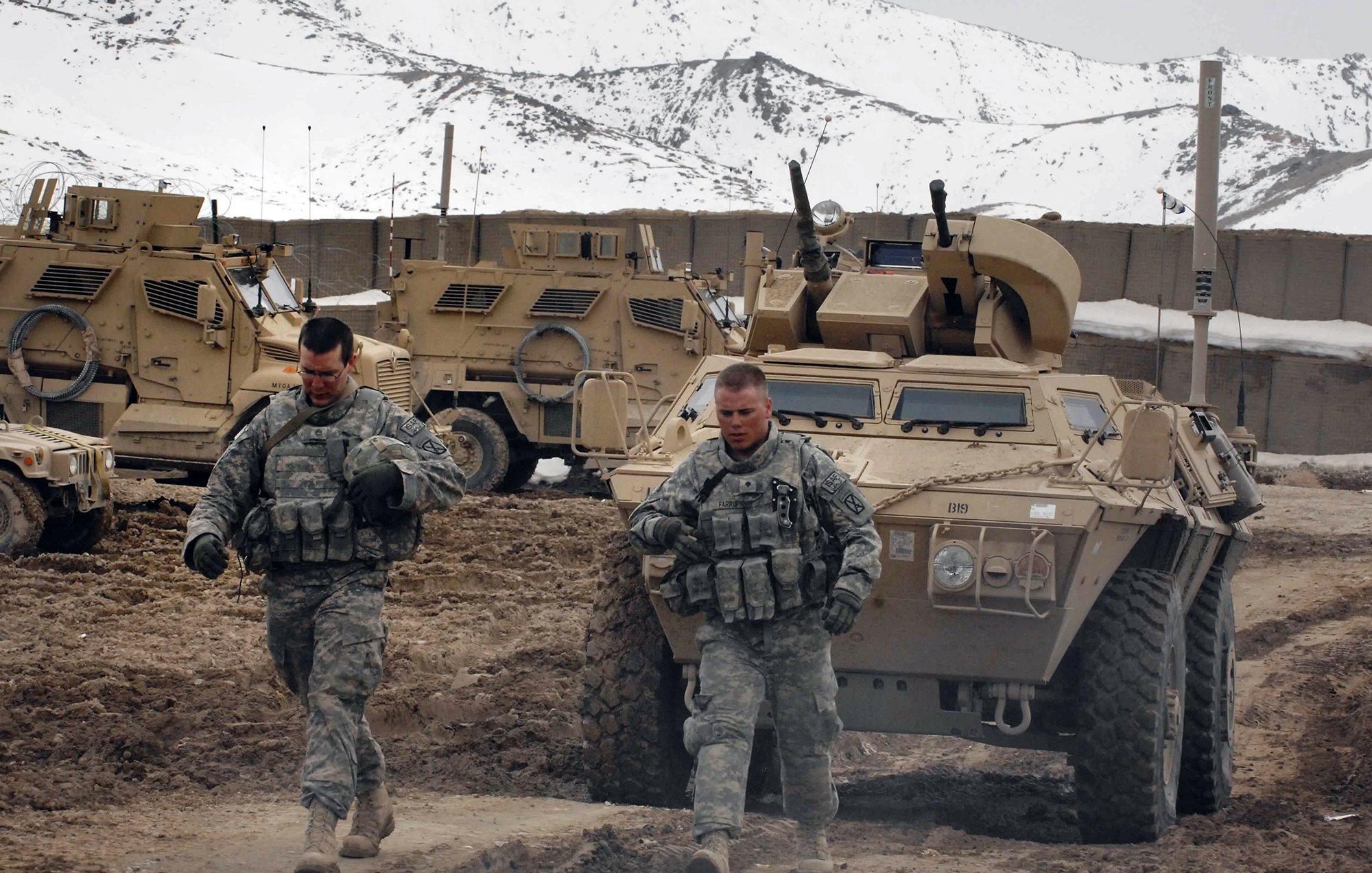 در این تصویر، دو سرباز در حال حرکت در یک زمین خاکی و سرد هستند. آنها لباس نظامی به تن دارند و در پس‌زمینه، چندین خودروی زرهی مشاهده می‌شود. کوه‌ها و برف در دوردست قابل مشاهده است که نشان‌دهنده شرایط جغرافیایی سخت منطقه است. سربازان به نظر می‌رسد که در حال انجام یک ماموریت یا گشت‌زنی هستند.