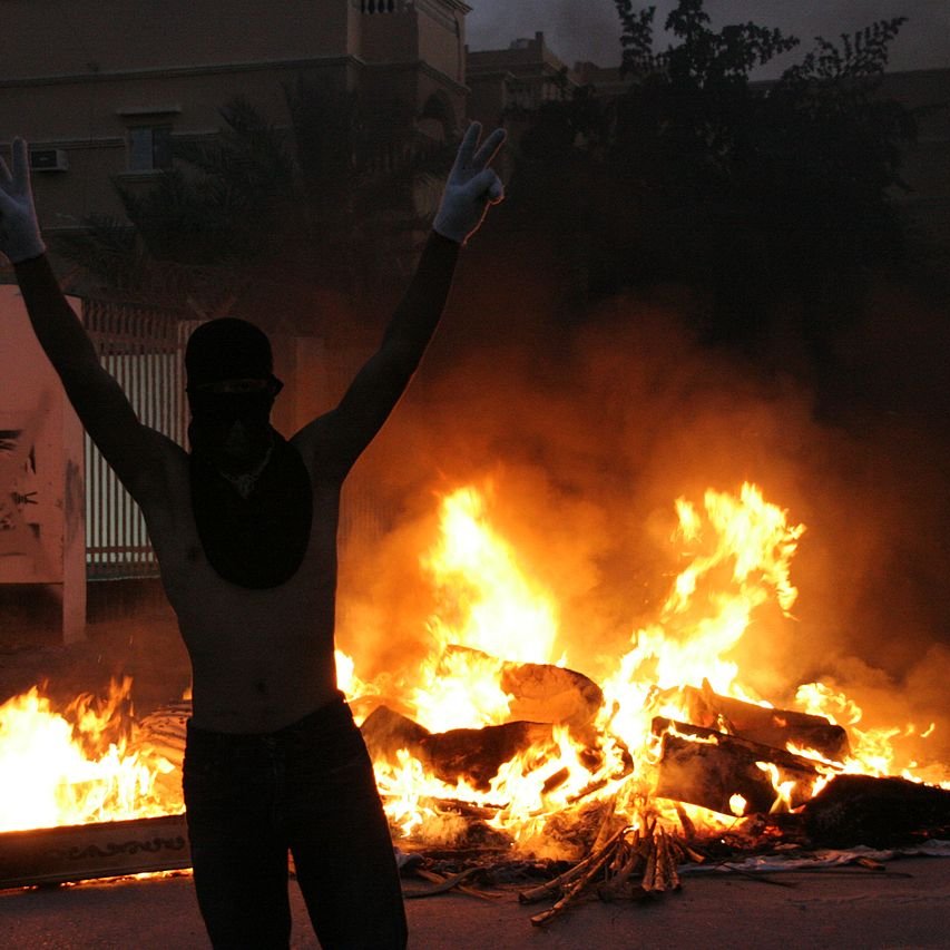 L'image montre une scène intense et dramatique. À l'avant-plan, une personne se tient avec les bras levés, formant un signe de victoire, tout en portant un masque qui couvre son visage. En arrière-plan, on peut voir des flammes vives qui s'élèvent, indiquant un feu, probablement une barricade ou des objets en feu. L'atmosphère est chargée d'émotion, évoquant un moment de protestation ou de turbulence. Les ombres des bâtiments environnants ajoutent à l'ambiance sombre de la scène.