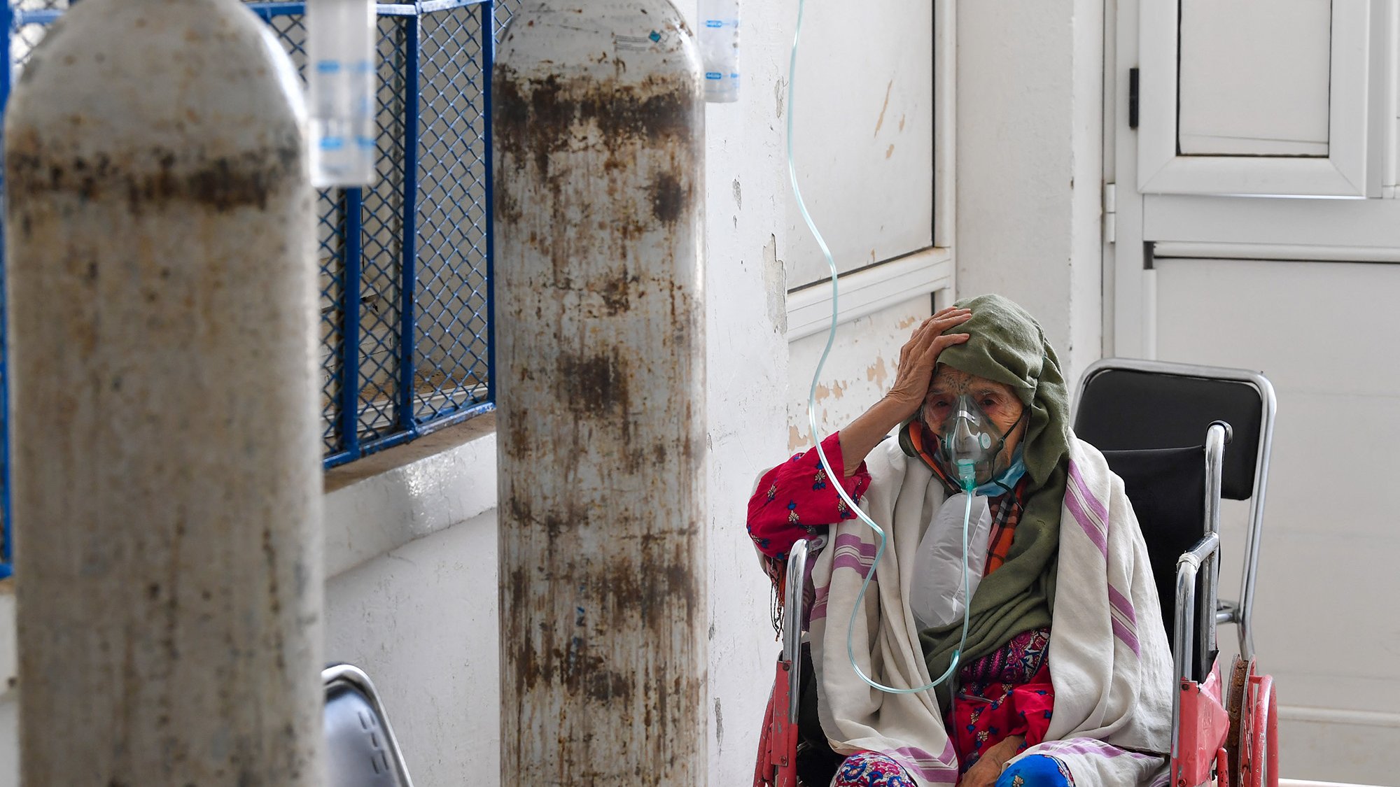 L'image montre une femme âgée assise dans un couloir d'hôpital. Elle porte un masque et une écharpe sur la tête. On peut voir des bouteilles d'oxygène à proximité, indiquant qu'elle reçoit des soins médicaux. L'environnement est simple, avec des murs blancs et des fenêtres qui laissent passer la lumière. L'expression de la femme suggère une certaine inquiétude ou fatigue.